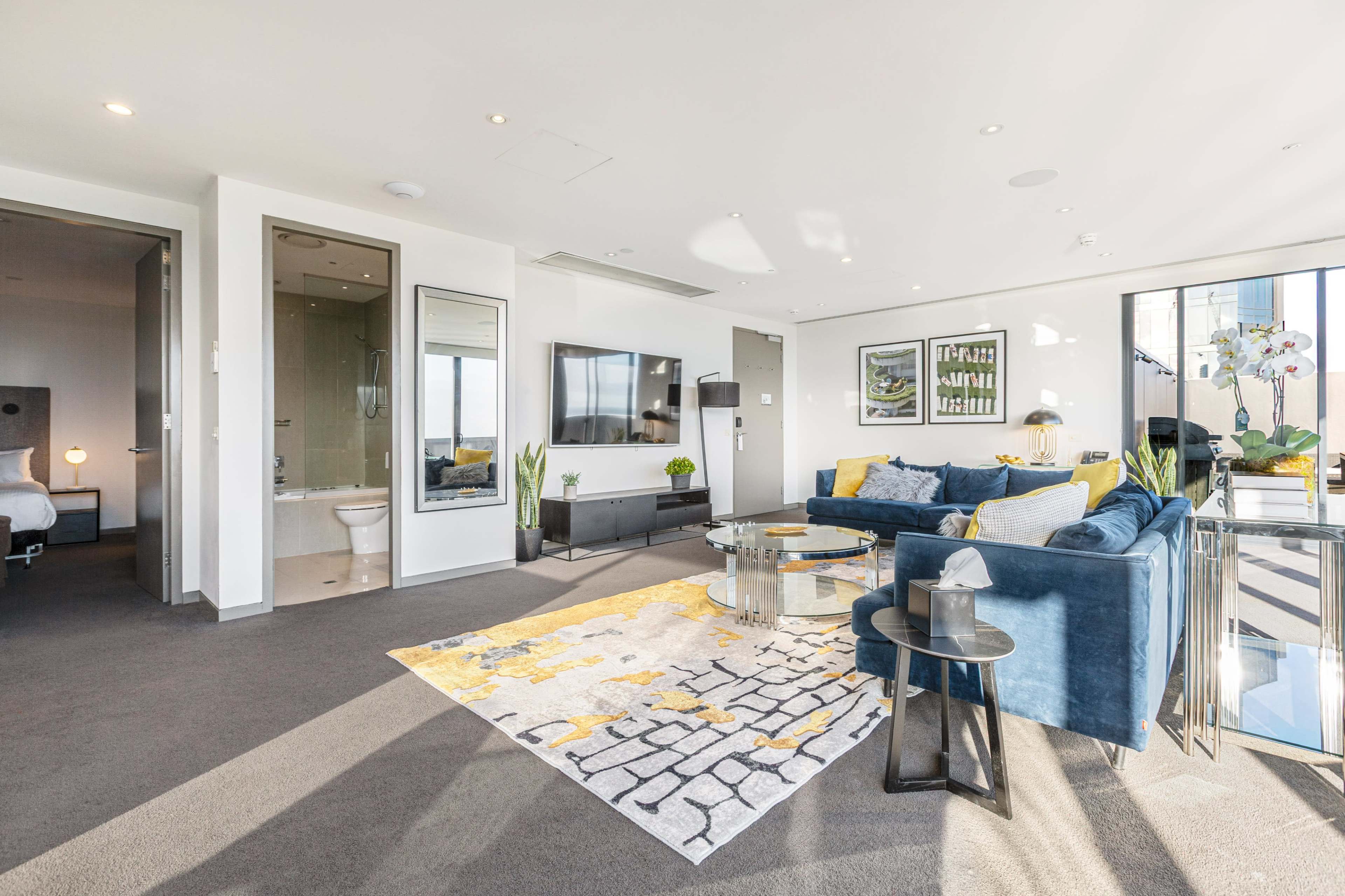 The image shows a bright, modern living room with a blue sectional sofa, a glass coffee table, and a view of a bedroom area through an open doorway.