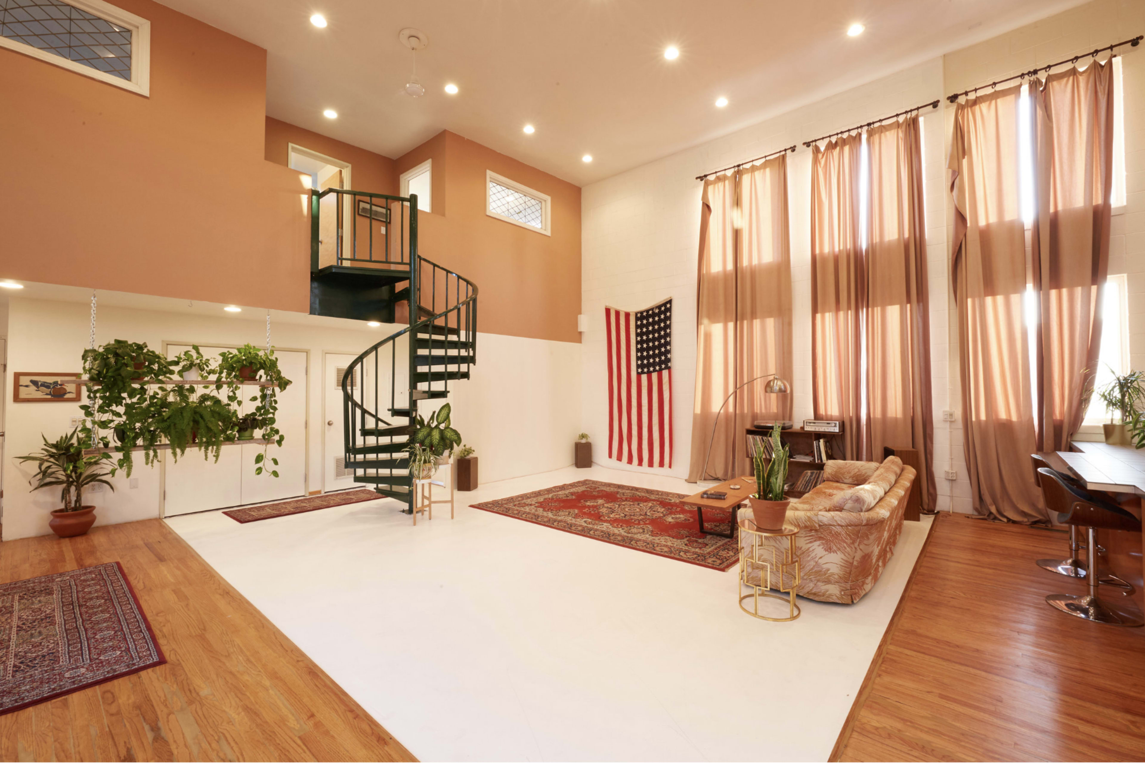 A spacious living area features a spiral staircase, large windows with draped curtains, an American flag, potted plants, and a mix of furniture on hardwood floors.