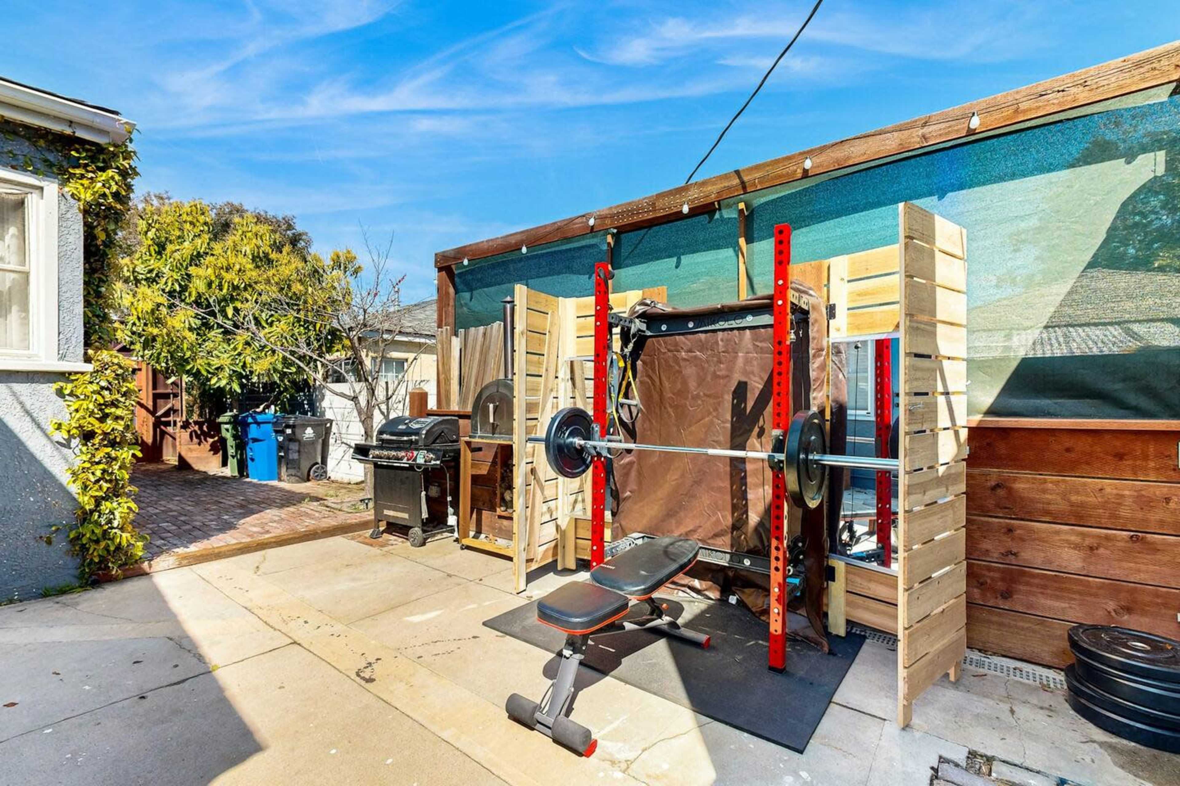 A backyard gym setup with a barbell rack and bench, accompanied by a grill and outdoor storage.