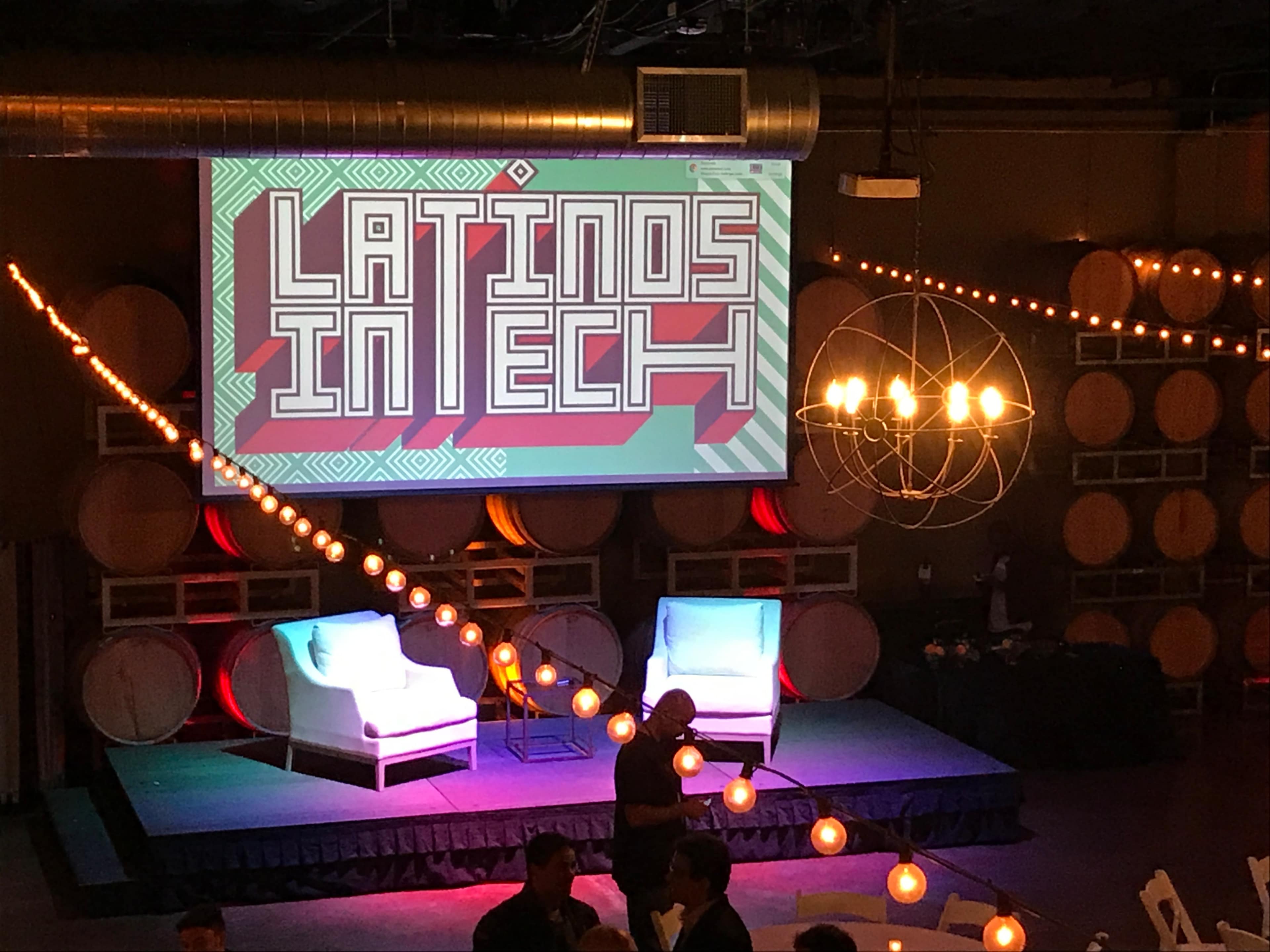 A stage setup featuring a large screen displaying "LATINOS IN TECH," flanked by two white chairs and decorated with string lights and barrels in the background.
