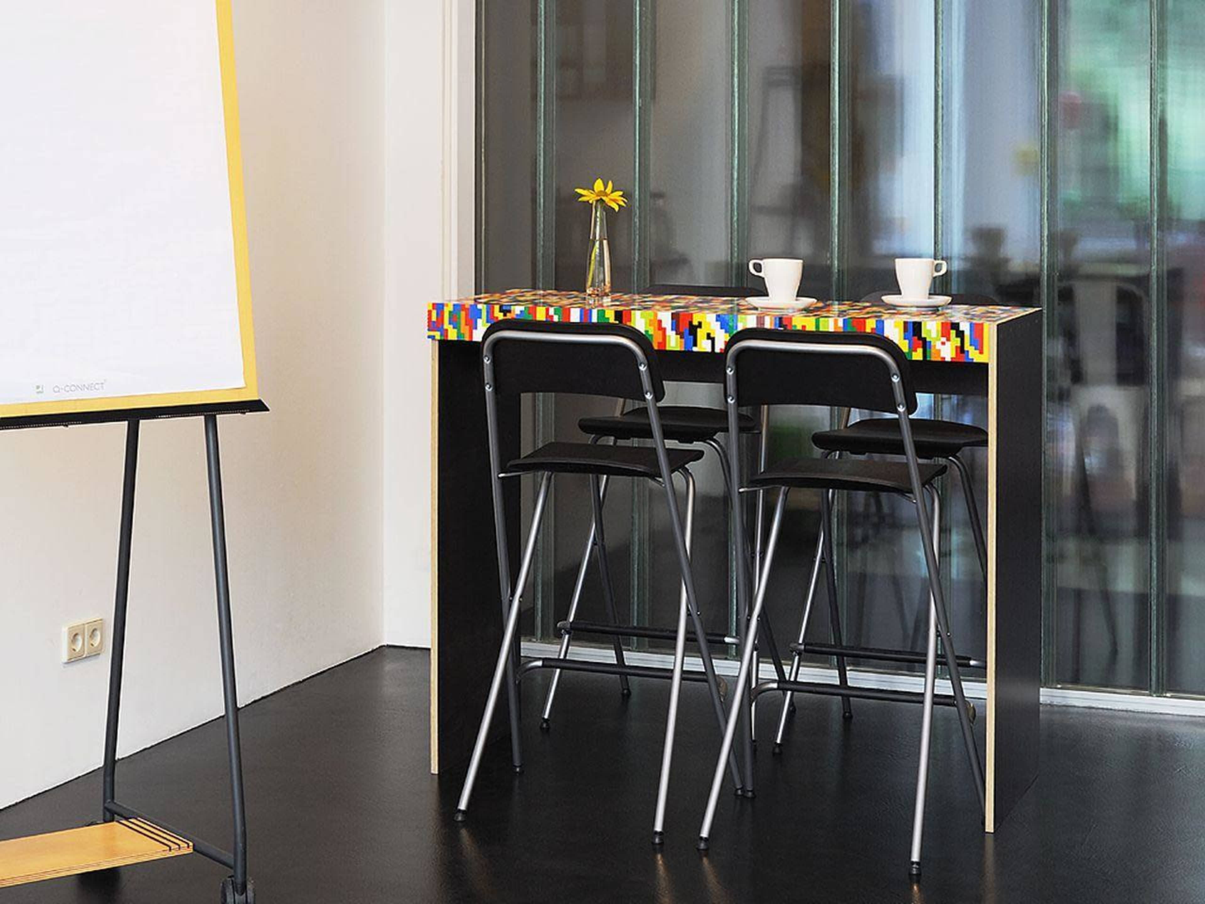 A black table with a colorful, patterned edge and two black folding chairs is positioned next to a glass wall, with a flower and two cups on the table.