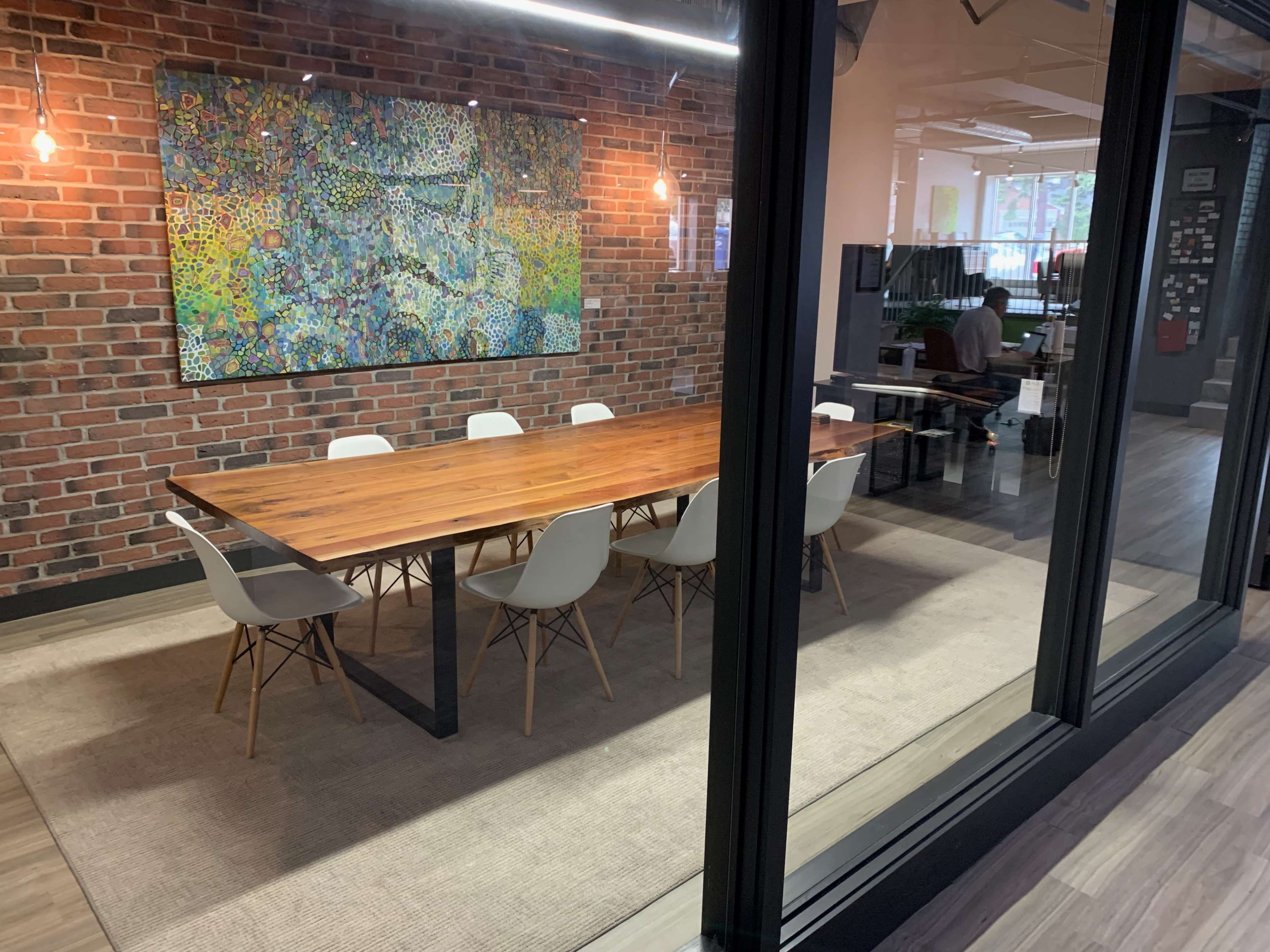 A large wooden dining table with white chairs is set in a glass-walled conference room featuring a colorful abstract painting on the brick wall.