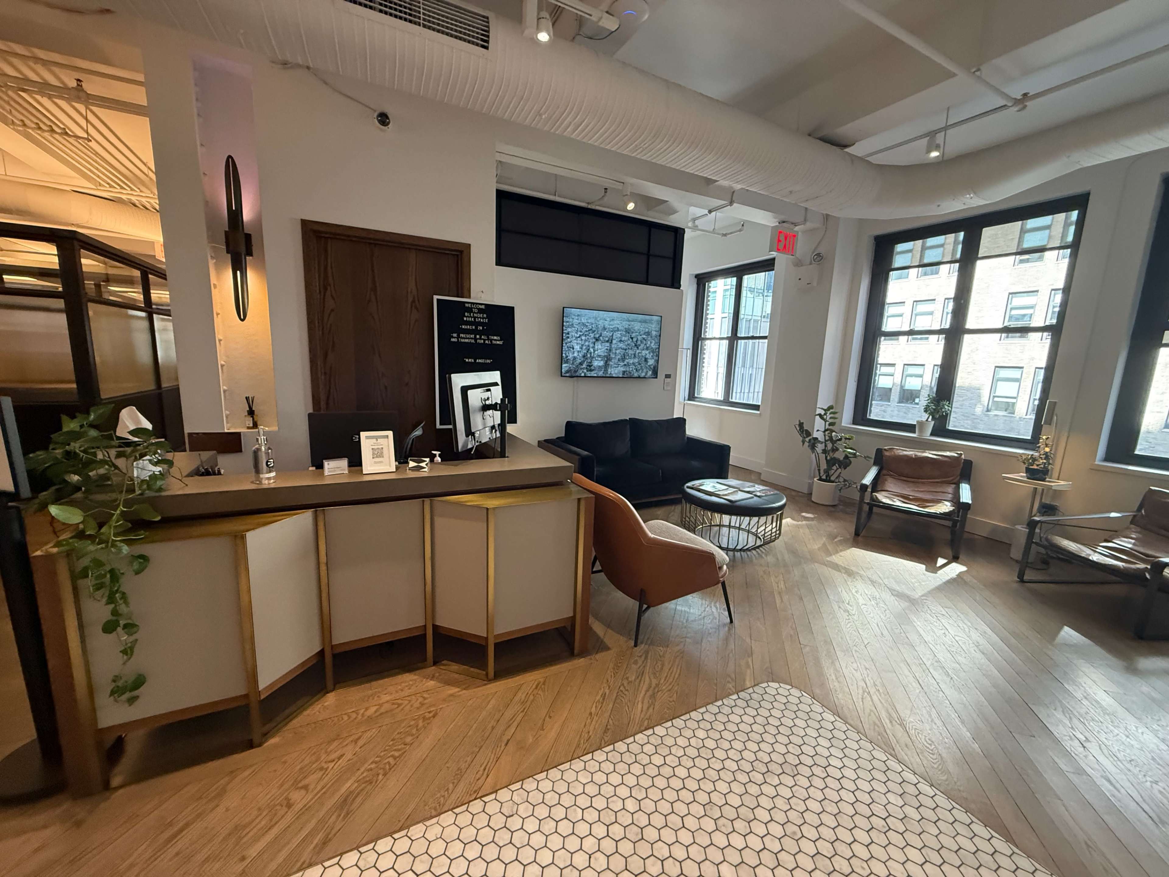 The image shows a modern, well-lit office reception area with wooden flooring, a desk, a black couch, and large windows.
