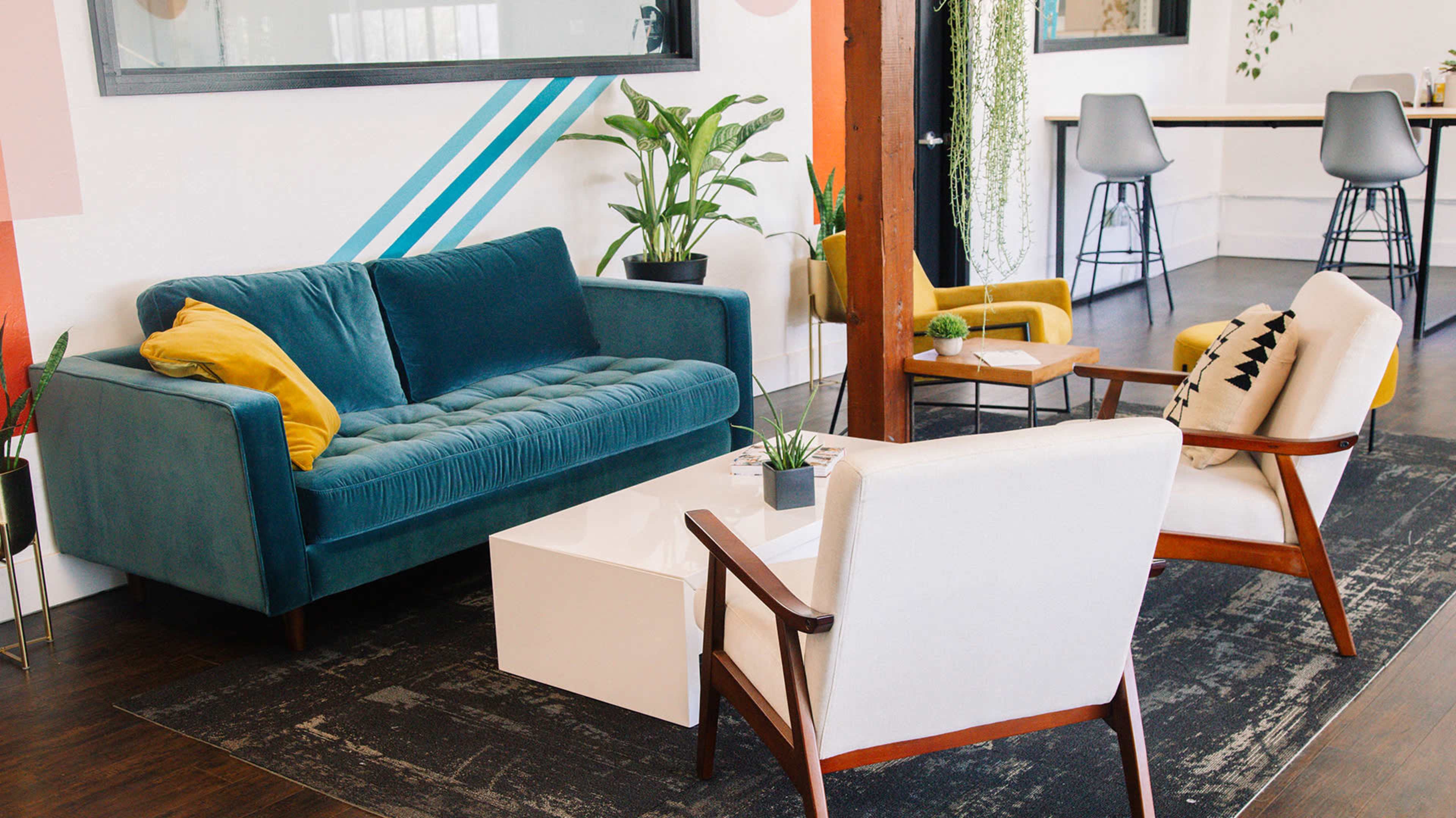 A modern seating area with a teal sofa and two light-colored armchairs arranged around a white coffee table on a patterned rug.