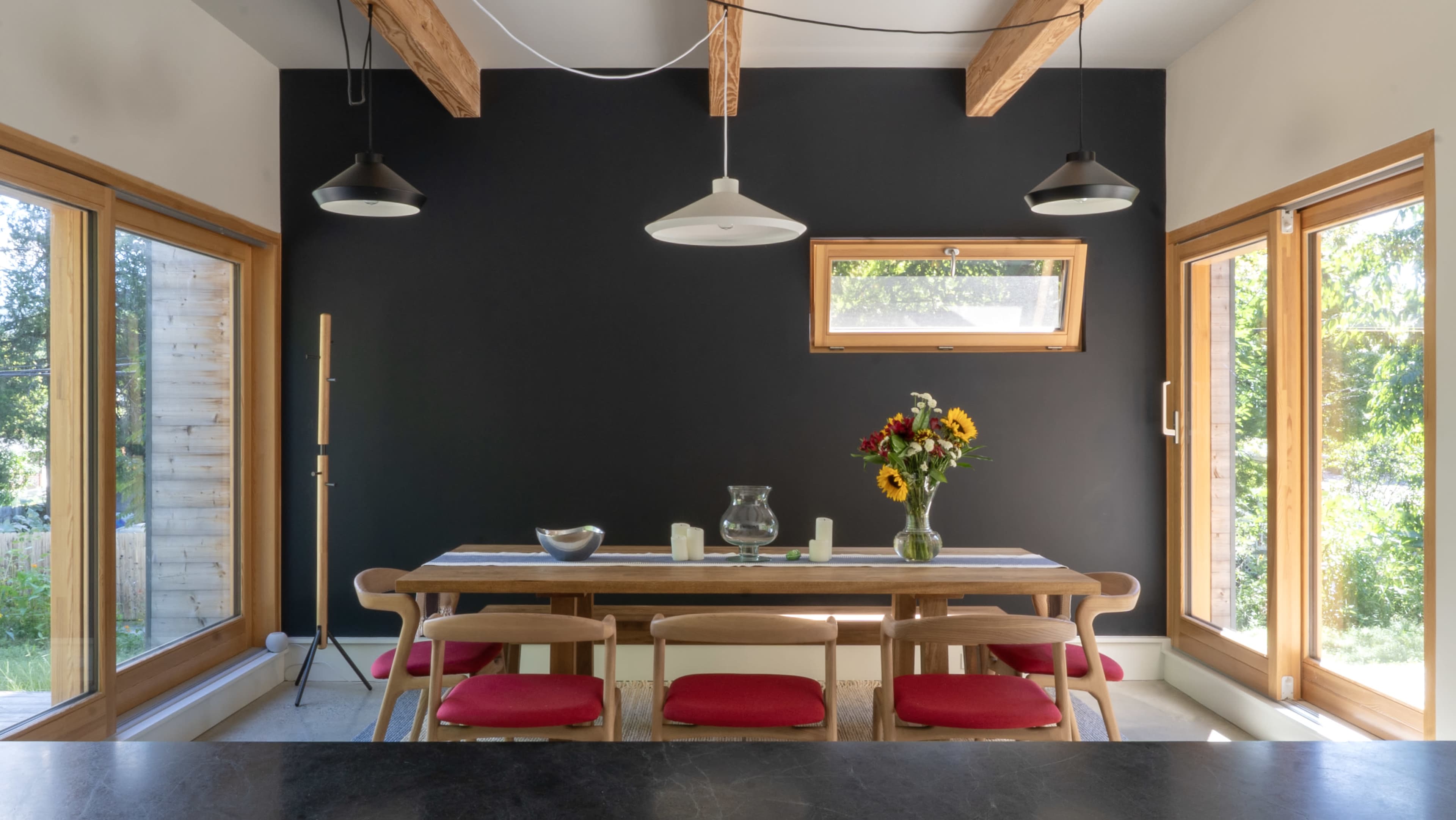 A dining area features a long wooden table surrounded by chairs, with large windows that let in natural light and a bouquet of sunflowers as a centerpiece.