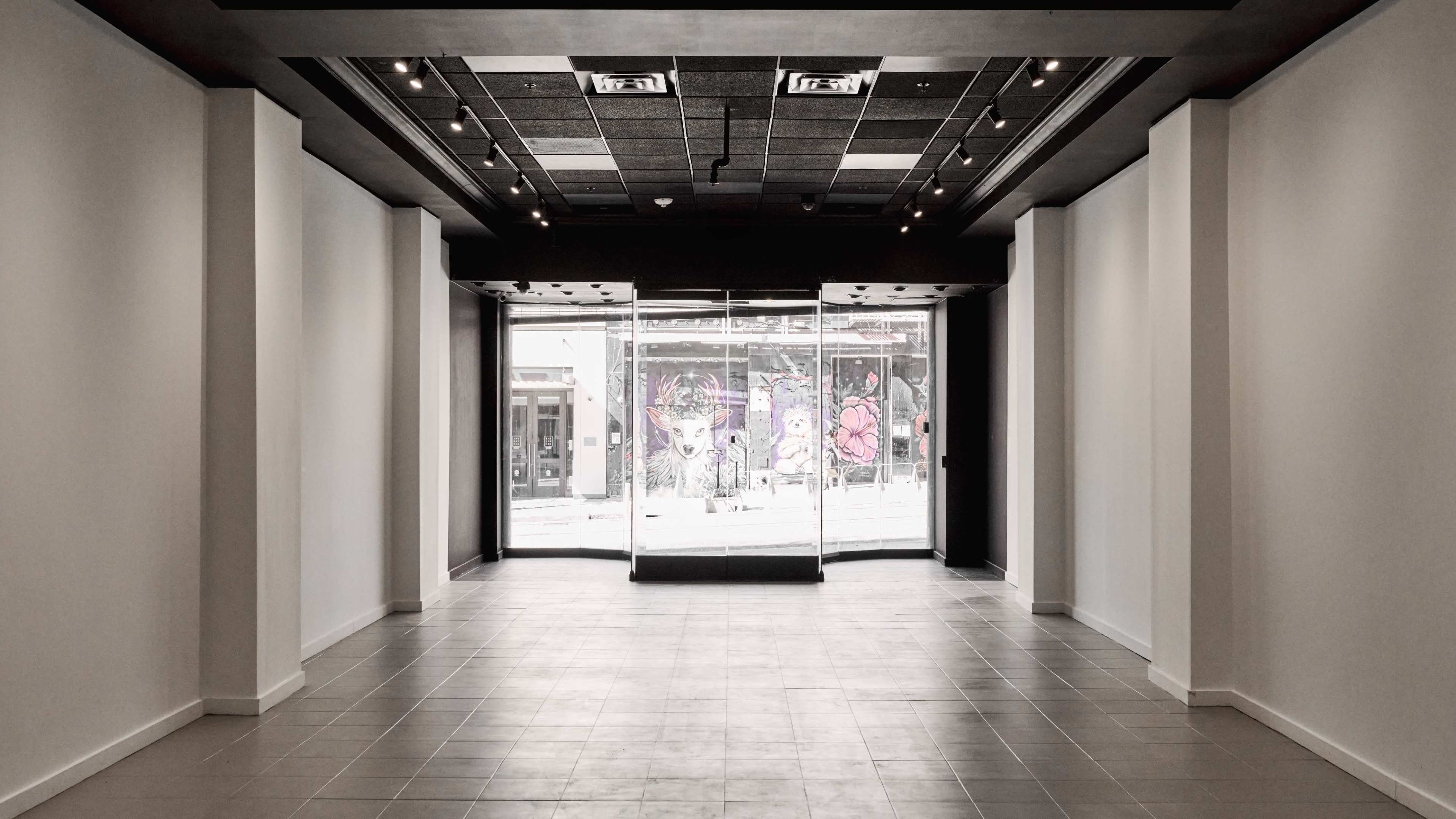 The image shows an empty interior of a retail space, featuring large glass doors that open to the street, with colorful murals visible outside.