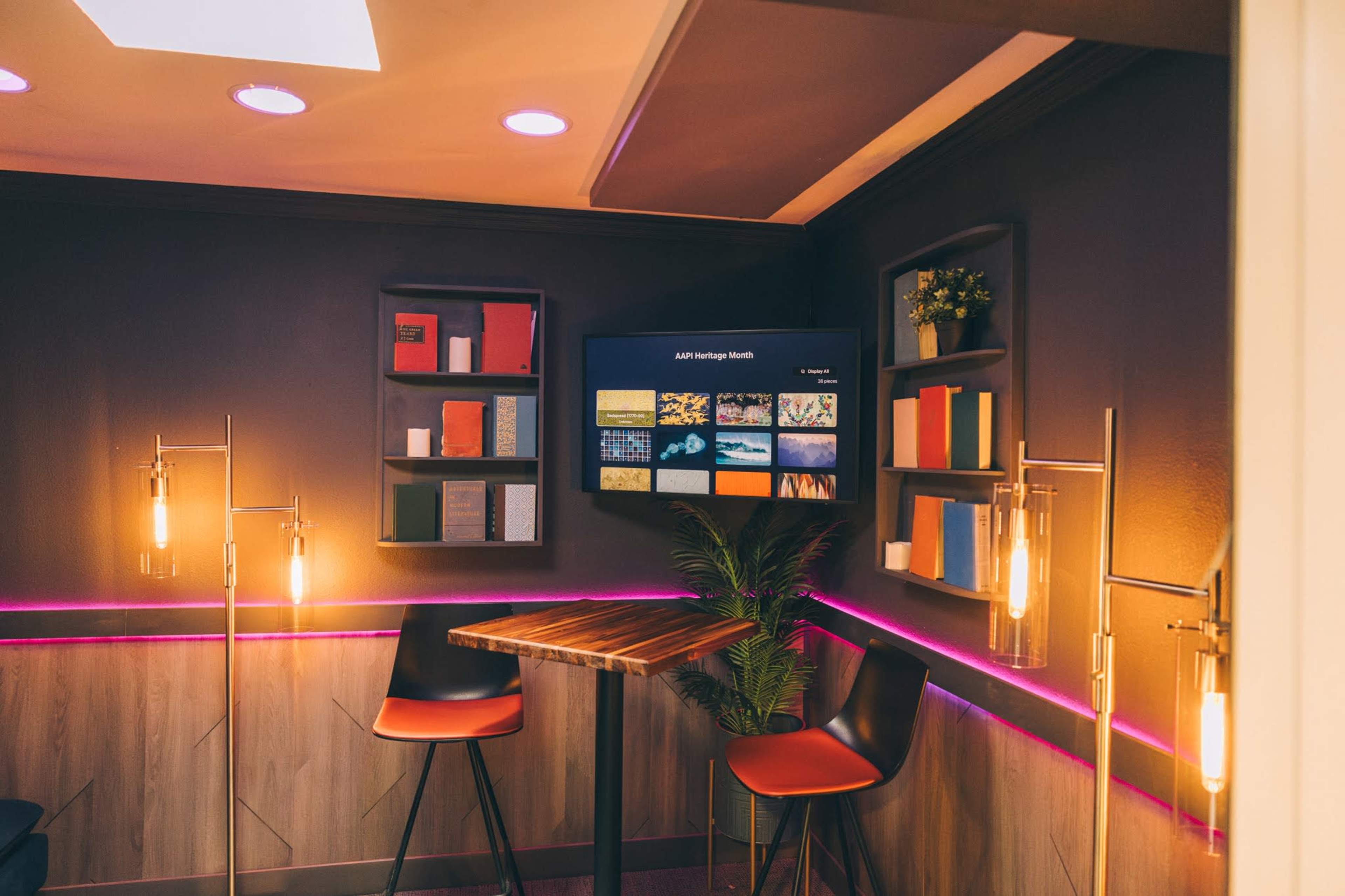 The image shows a contemporary corner space featuring a wooden table, sleek chairs, and shelves with decorative books and a screen displaying various media.