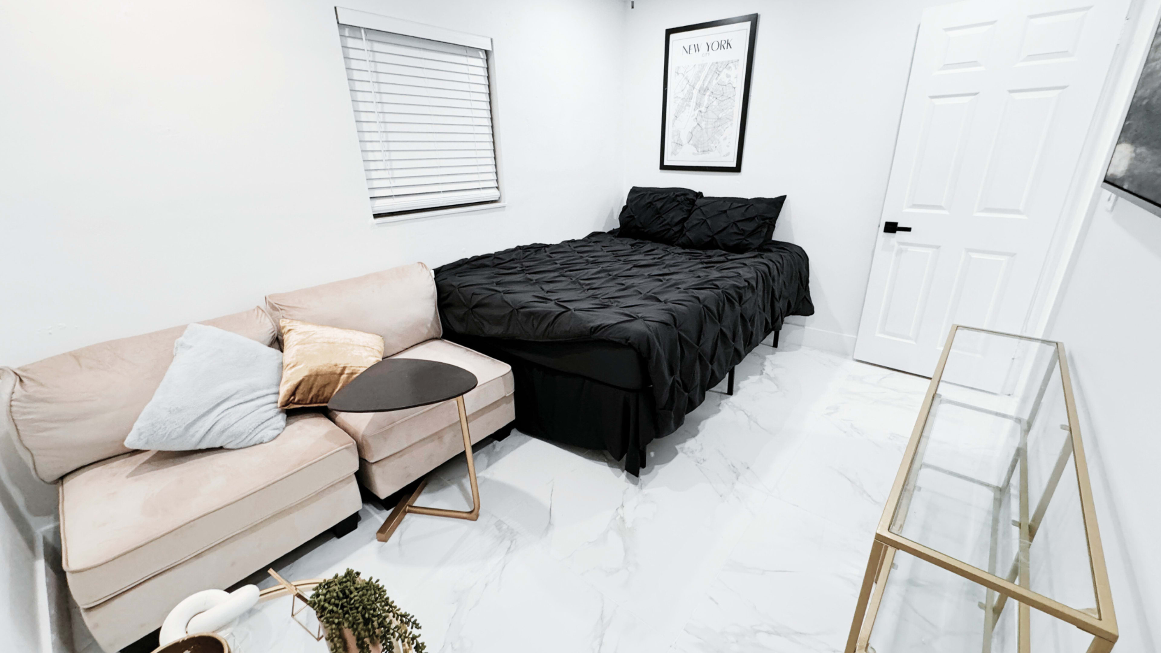 A small room with a bed covered in a black bedspread, a beige sectional couch, a gold-accented side table, and a glass console table against a white wall.
