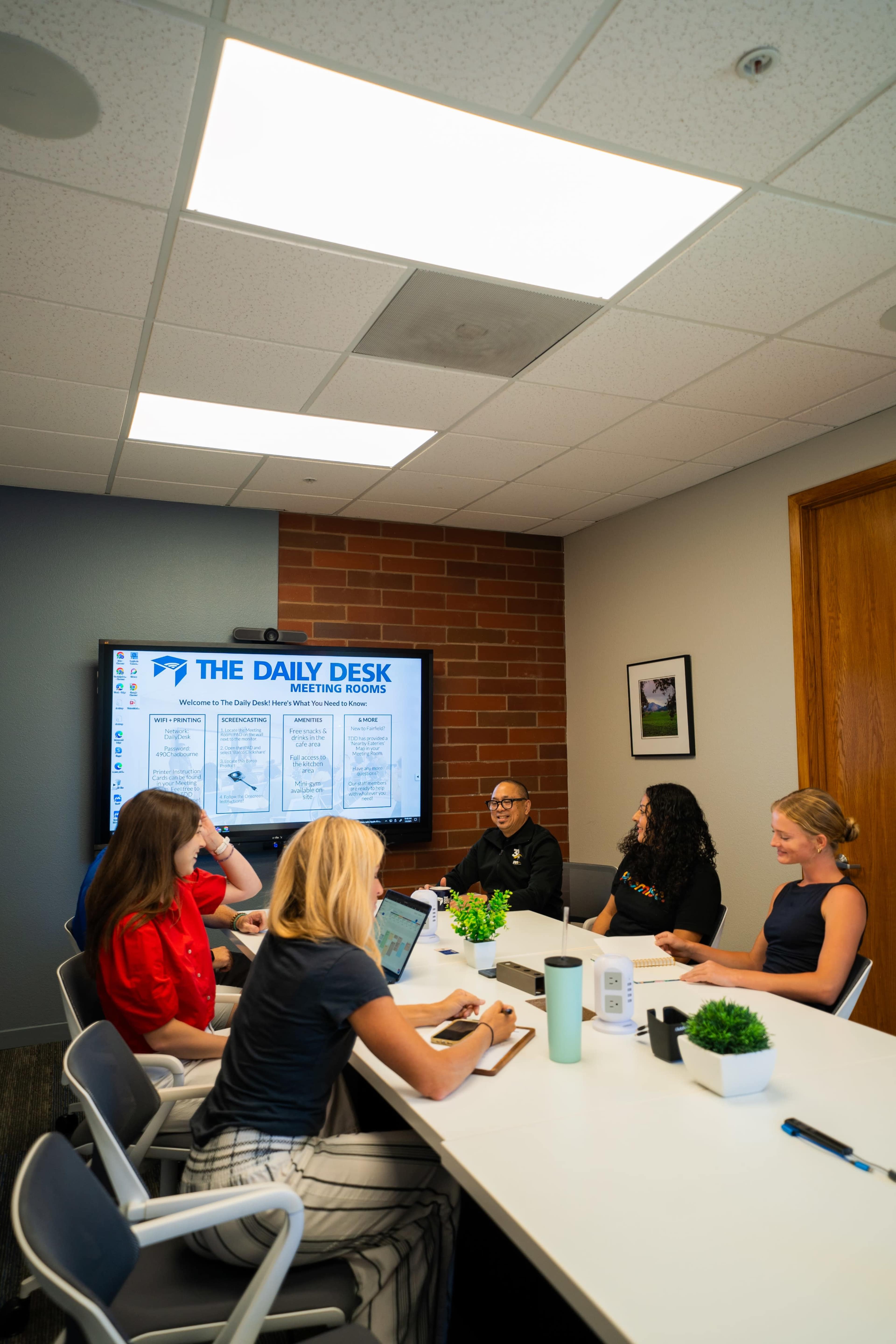 A group of six people sits around a conference table in a meeting room, with a television displaying information on the wall.