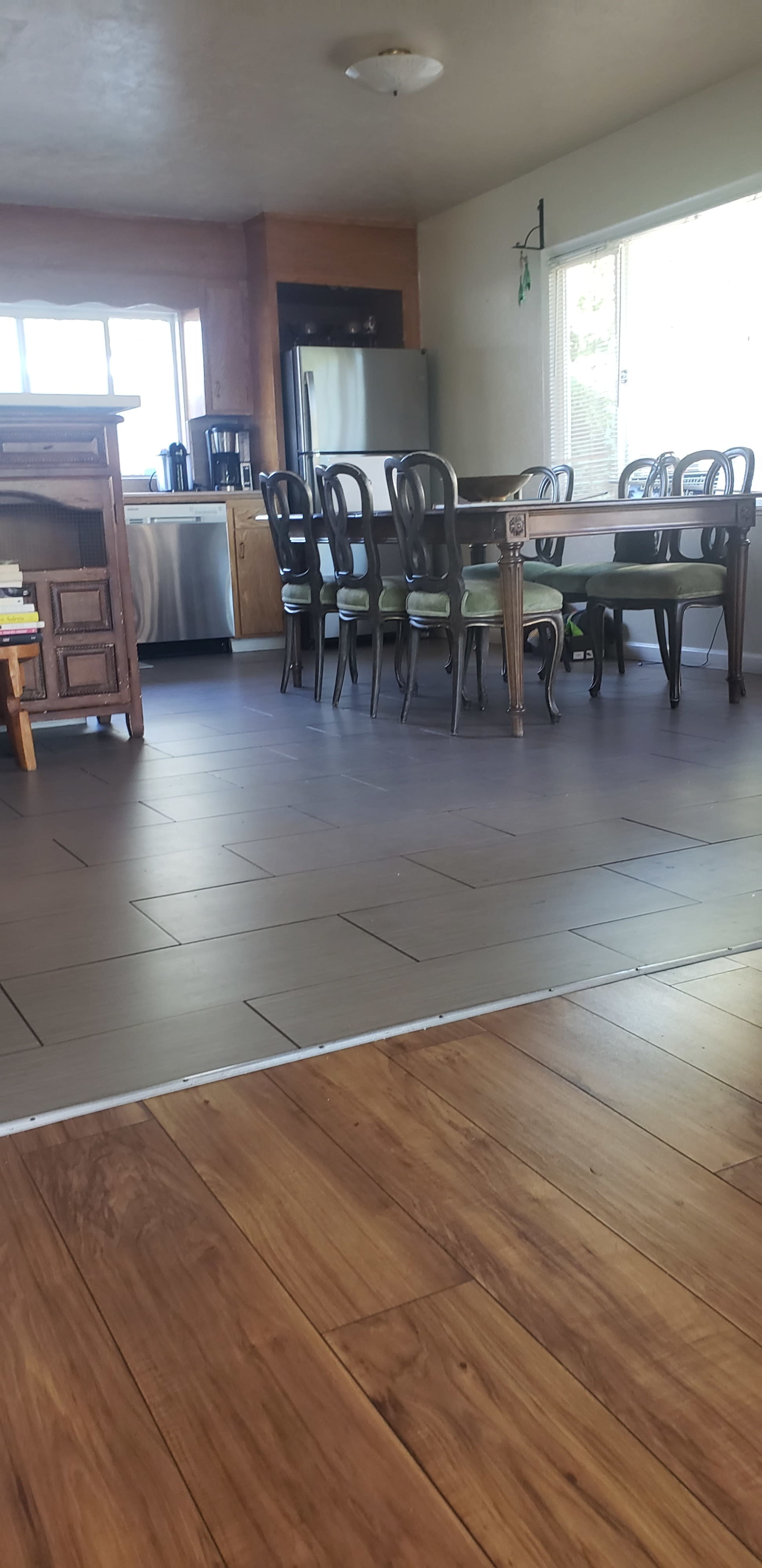 The image shows a dining area with a table and chairs, adjacent to a kitchen featuring appliances and a window that allows natural light.