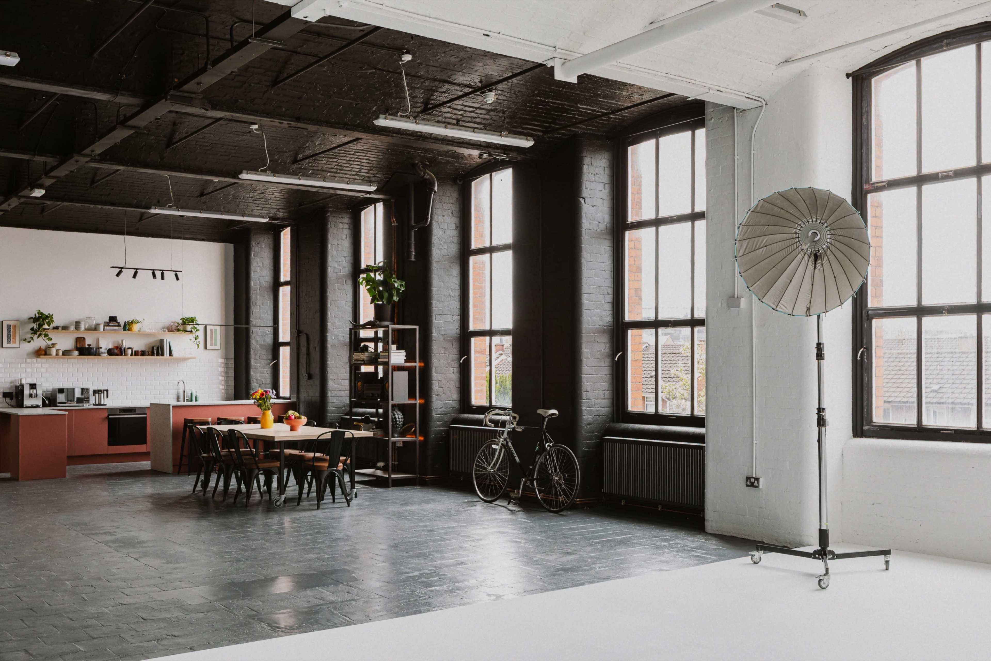 The image shows a spacious, modern studio with large windows, a kitchen area, a dining table, and a bicycle leaning against a rack.