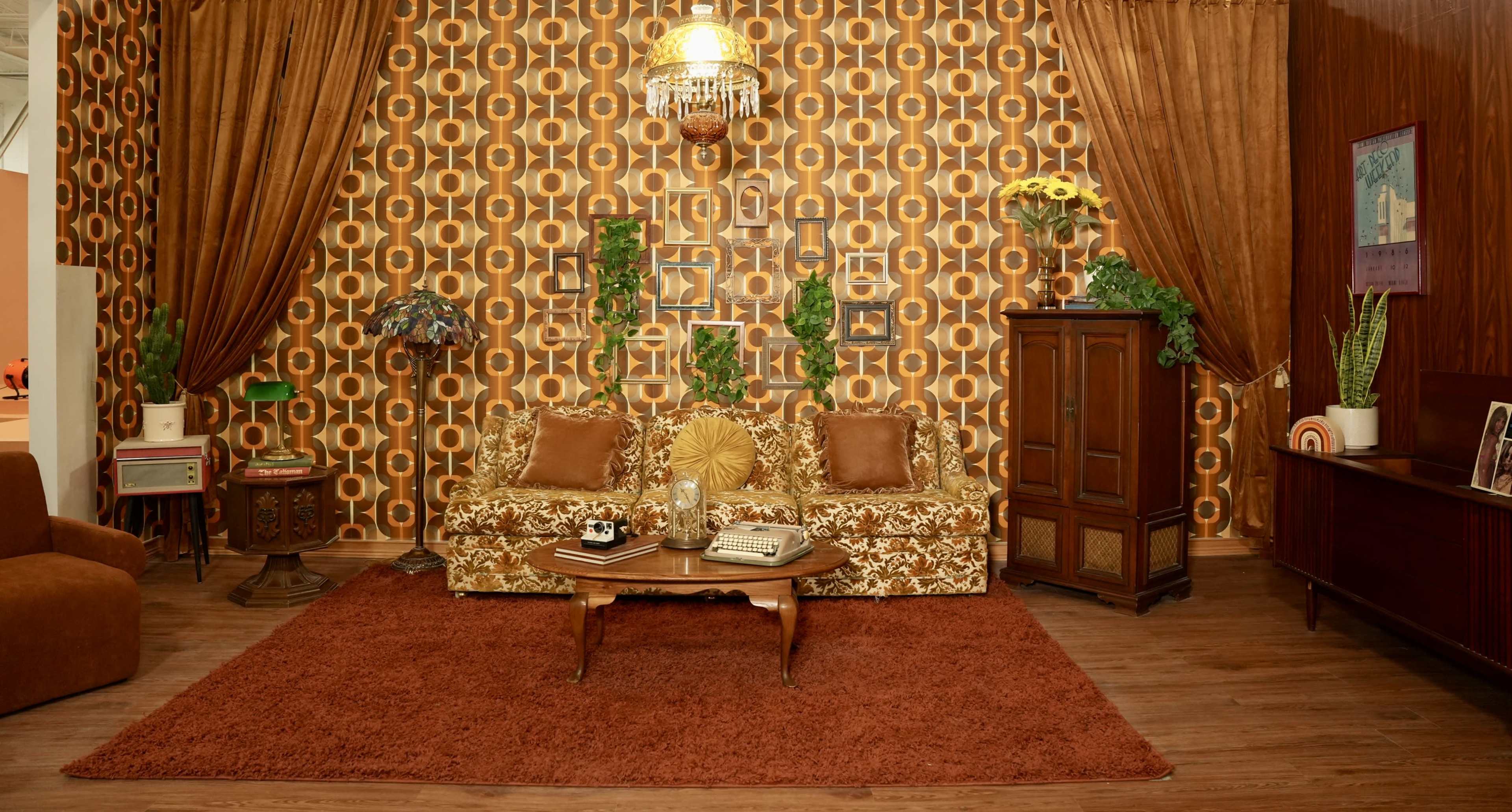 A vintage living room with patterned wallpaper, a floral sofa, a wooden cabinet, and a round coffee table on a carpet.