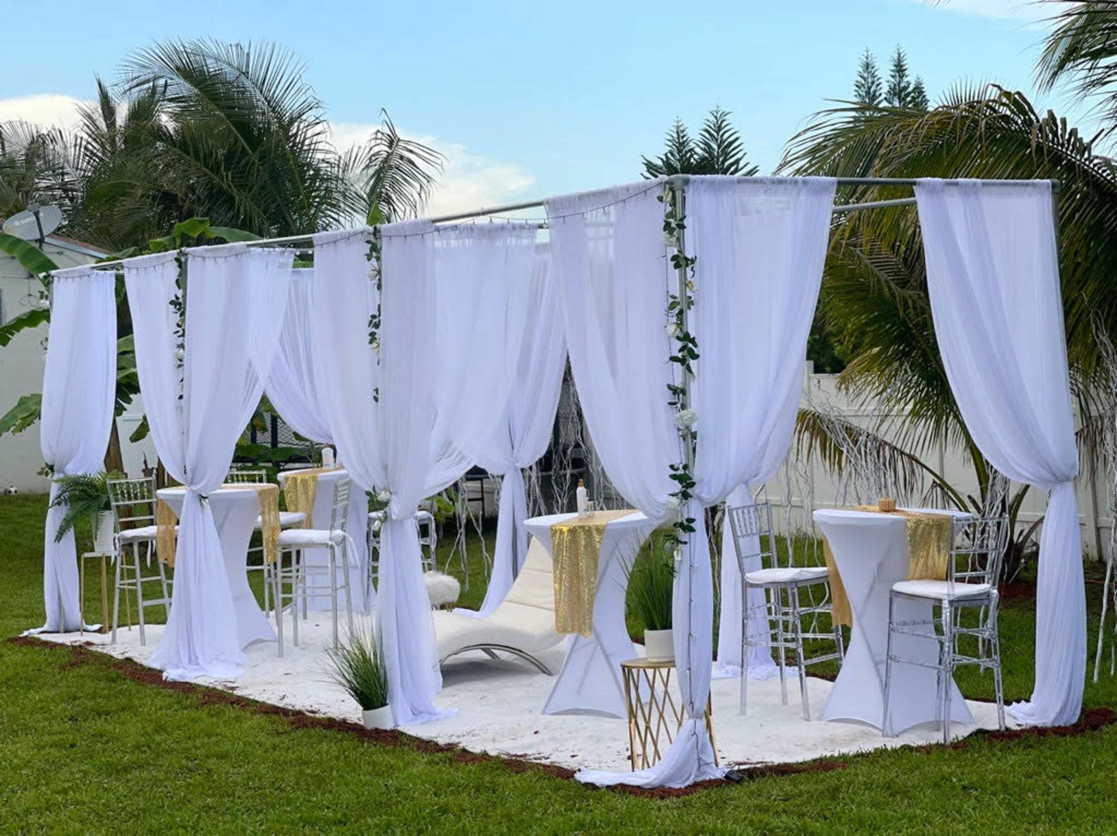 The image shows a decorated outdoor space with white sheer curtains, tables, and seating arranged in a garden setting.