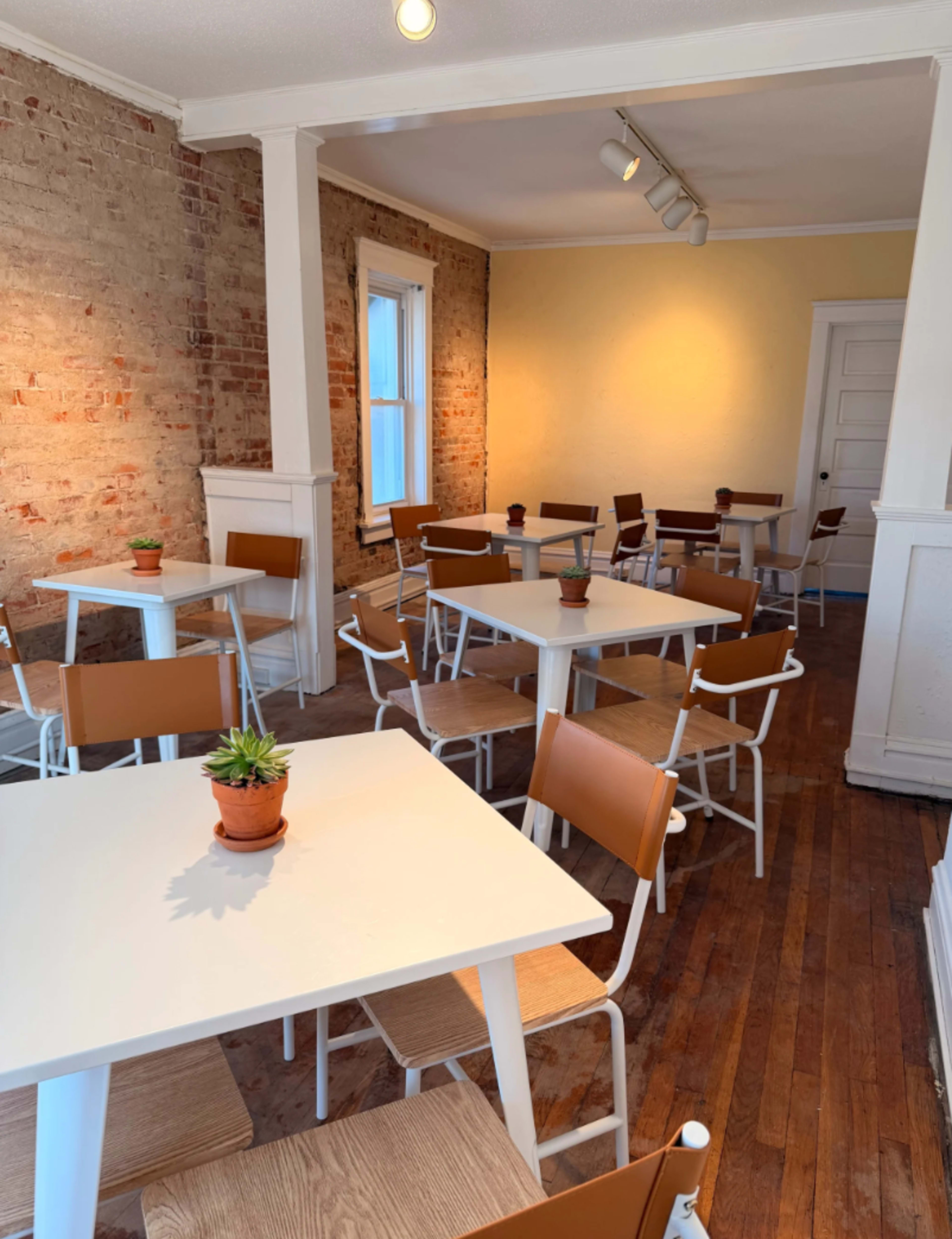 The image shows a bright, minimalistic cafe with white tables and chairs arranged neatly on a wooden floor, featuring potted plants on each table and exposed brick walls.