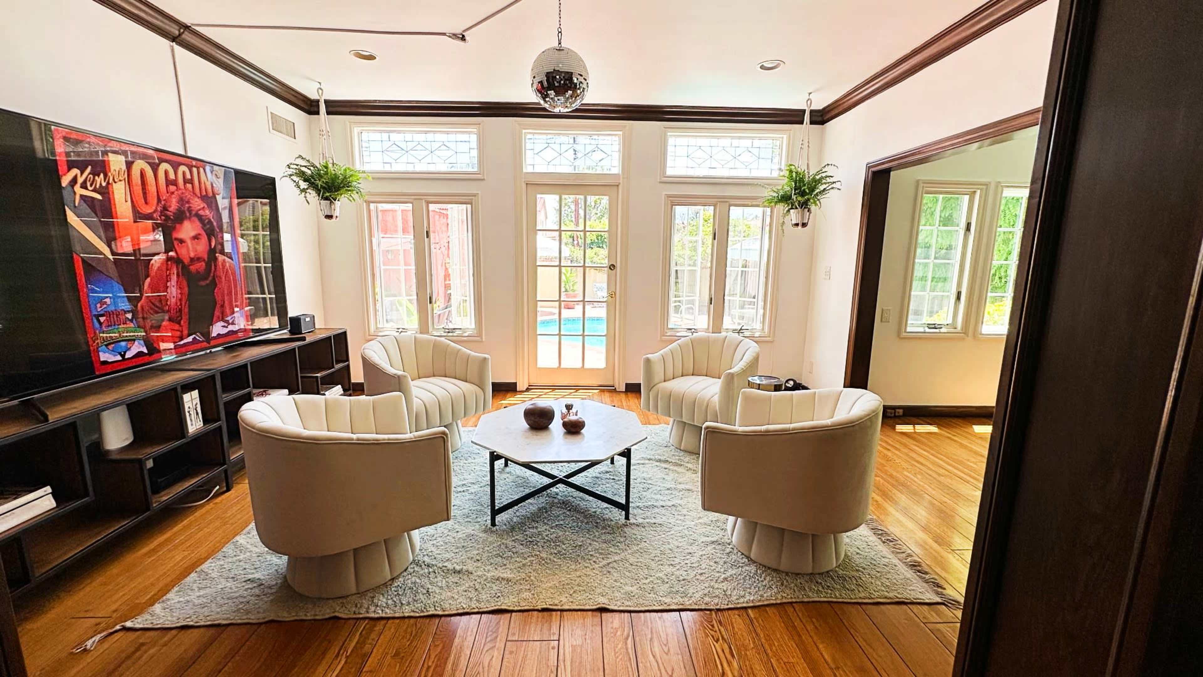 A living room featuring four white chairs arranged around a black coffee table on a light rug, with a large television mounted on the wall and doors leading to a pool area in the background.