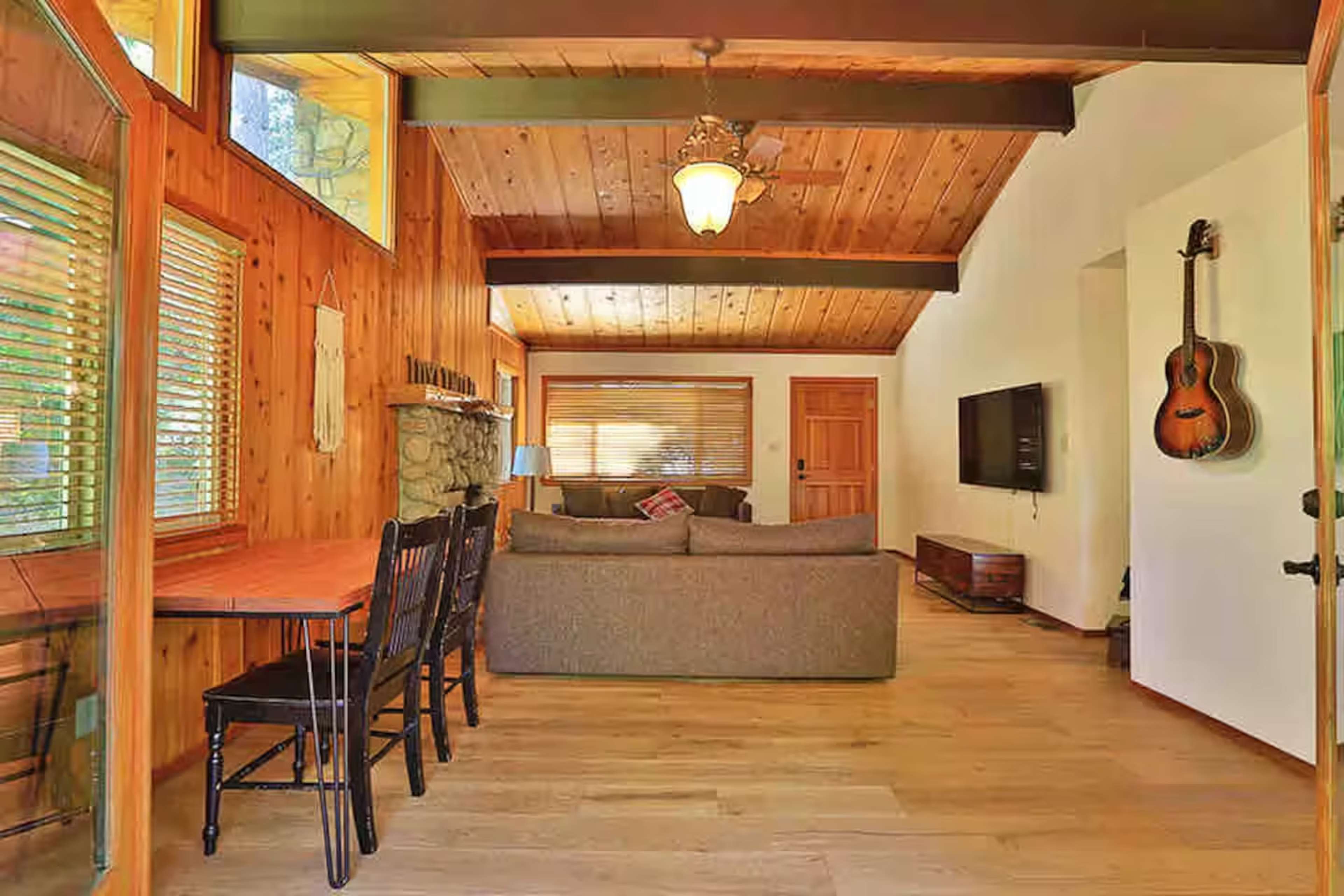 The image shows a rustic living area with wooden walls, a comfortable gray sofa, a dining table with chairs, and a television mounted on the wall.
