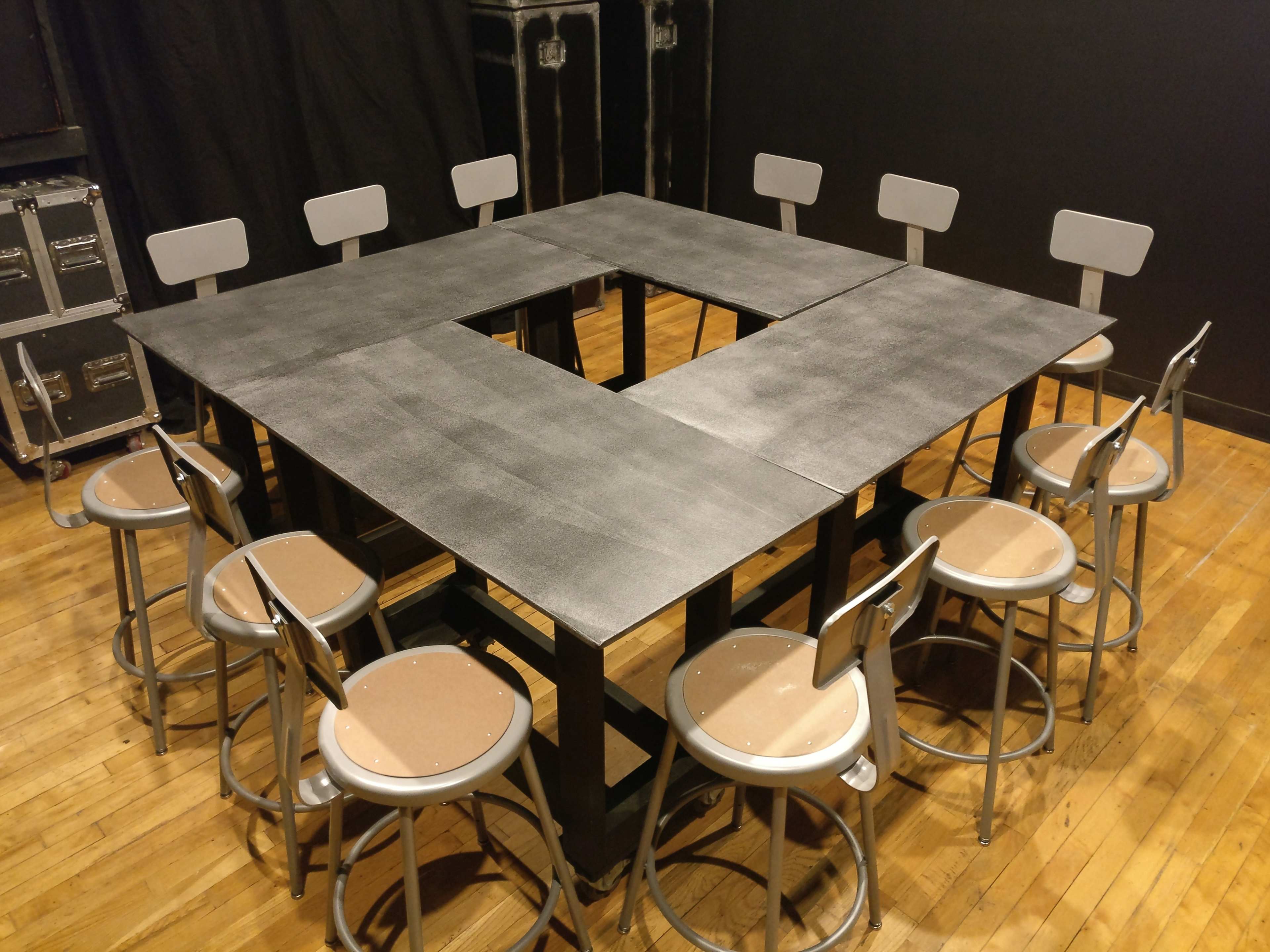 A square table with a cutout in the center is surrounded by eight metal stools on a wooden floor.