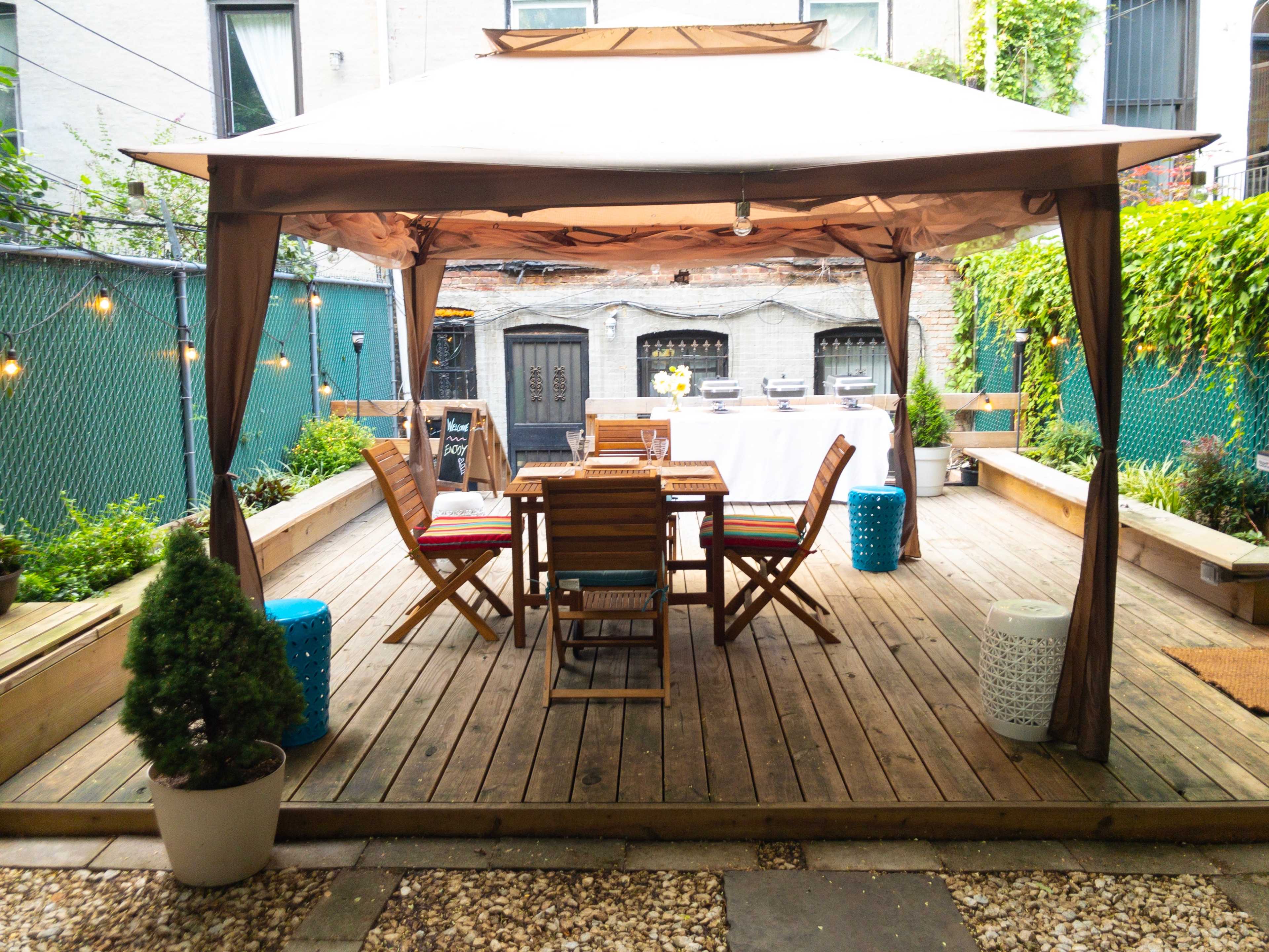 A wooden deck features a gazebo with a table and chairs surrounded by greenery and decorative accents.