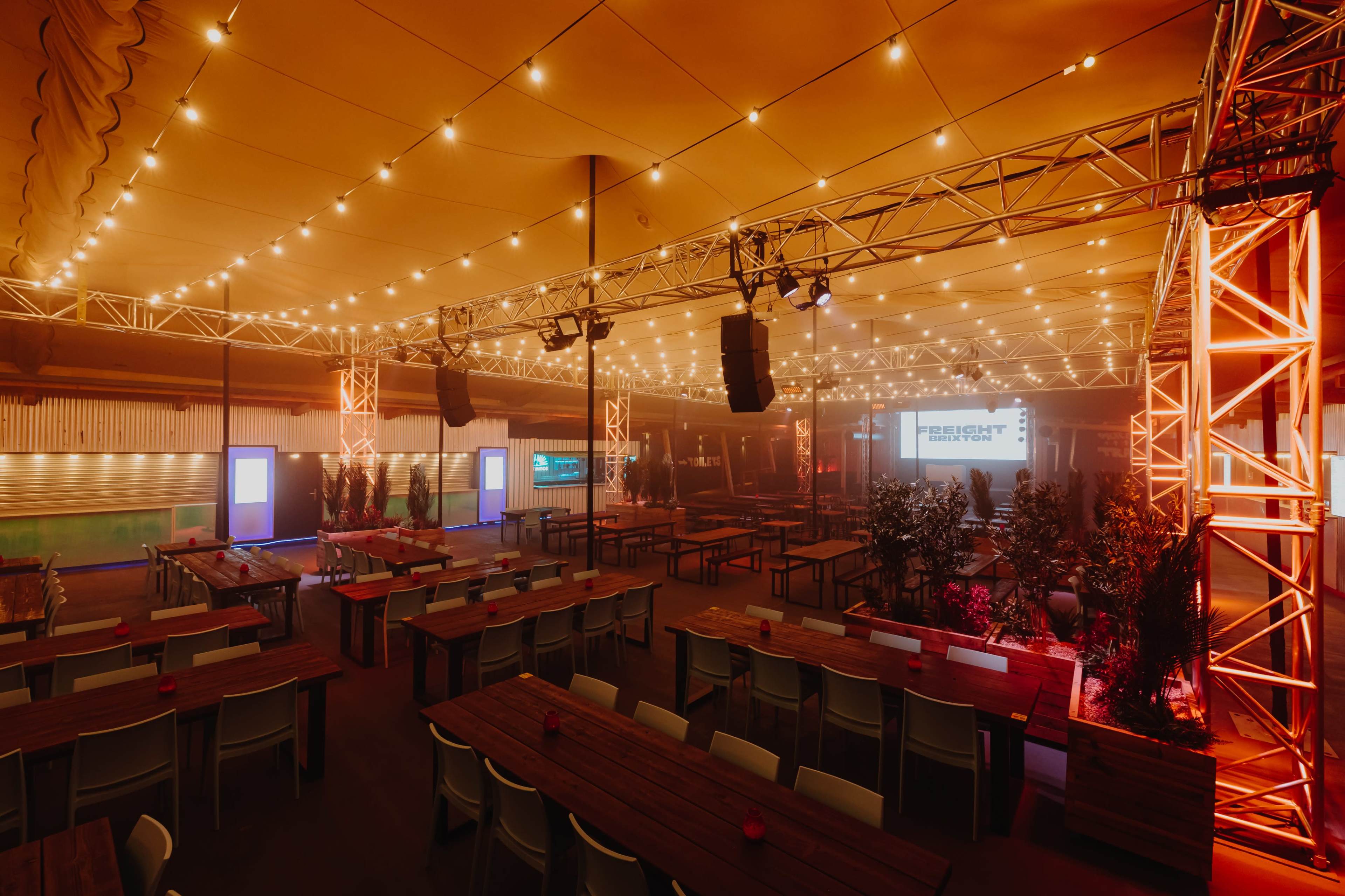 The image shows a large indoor event space with rows of wooden tables, string lights overhead, and a stage area at one end, illuminated with colorful lights.