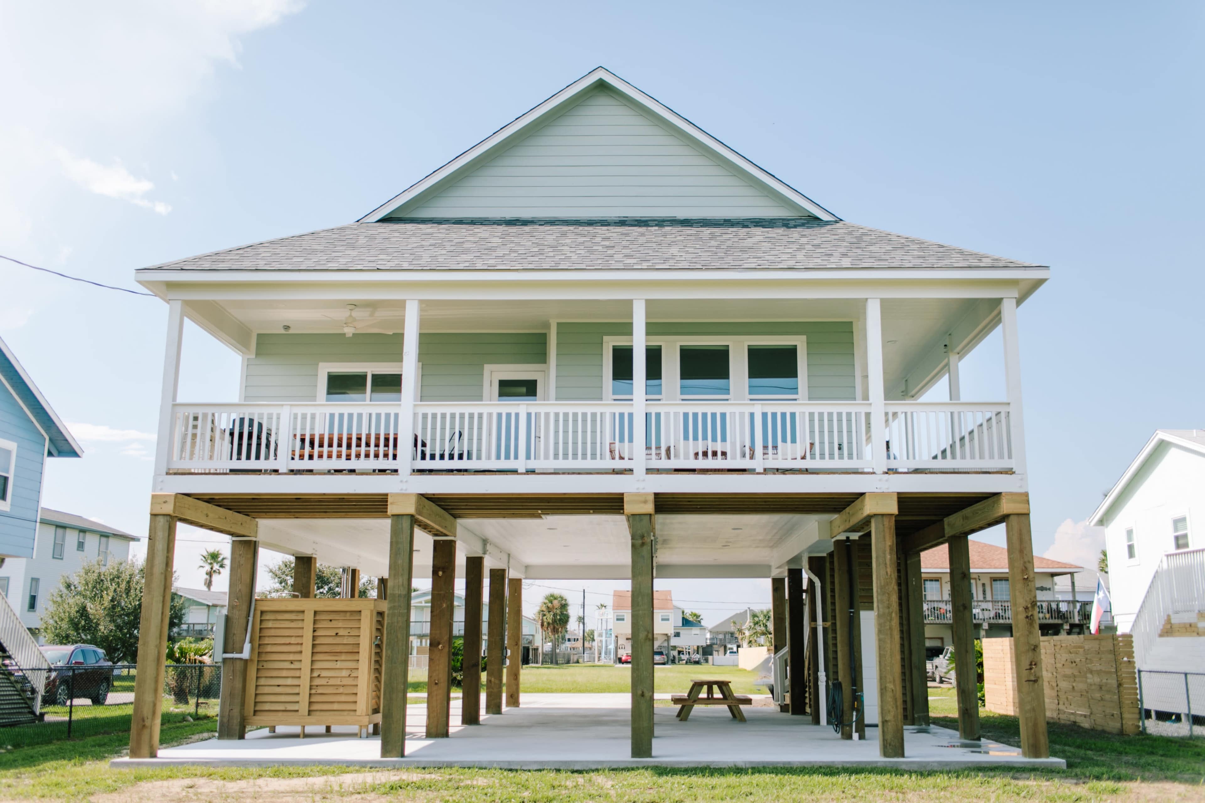 A two-story house with a light blue exterior and a spacious deck stands on stilts above a concrete area, surrounded by grass and nearby homes.