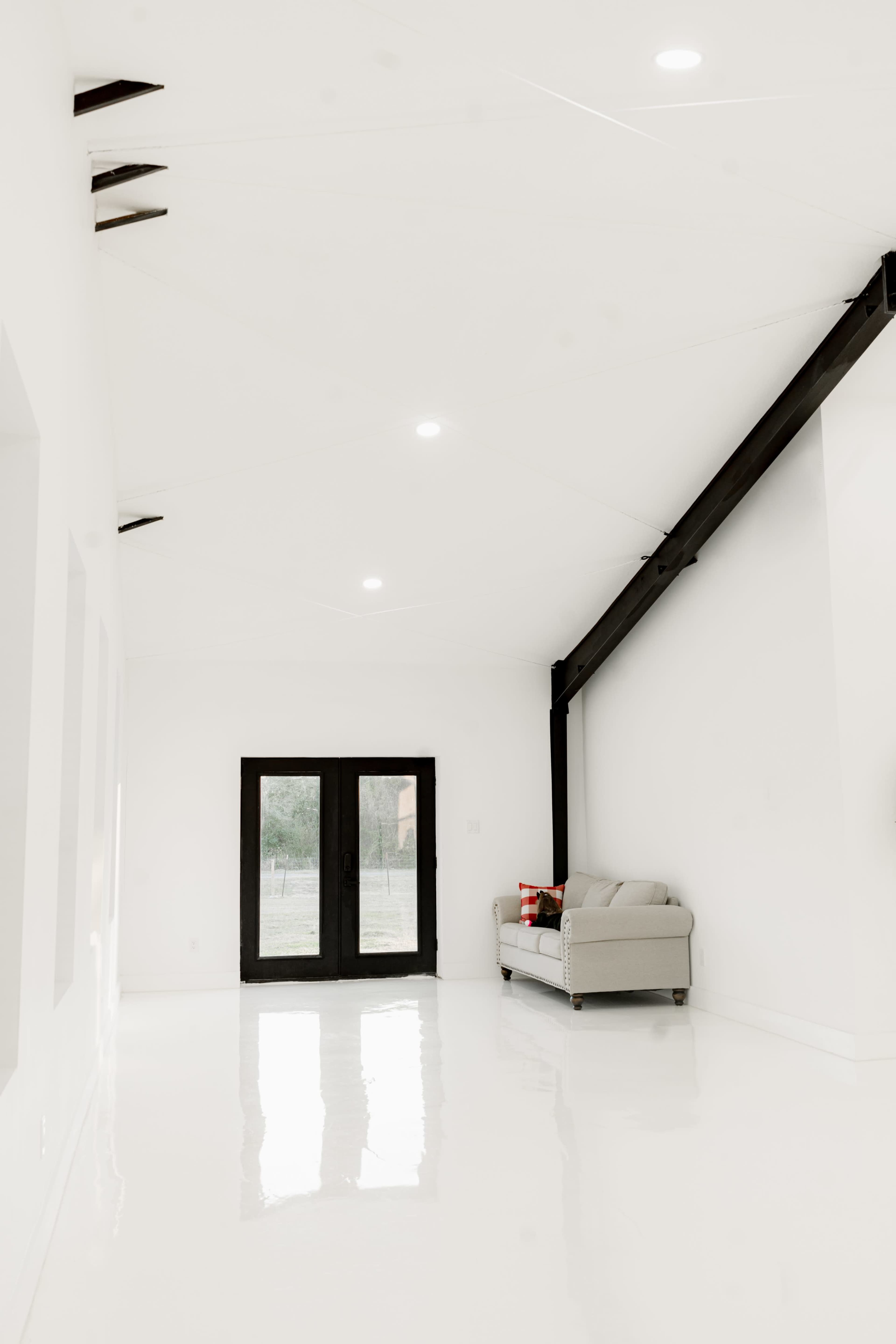 The image shows a spacious, bright room with white walls, polished floors, and a gray sofa near a set of black double doors leading to an outdoor view.