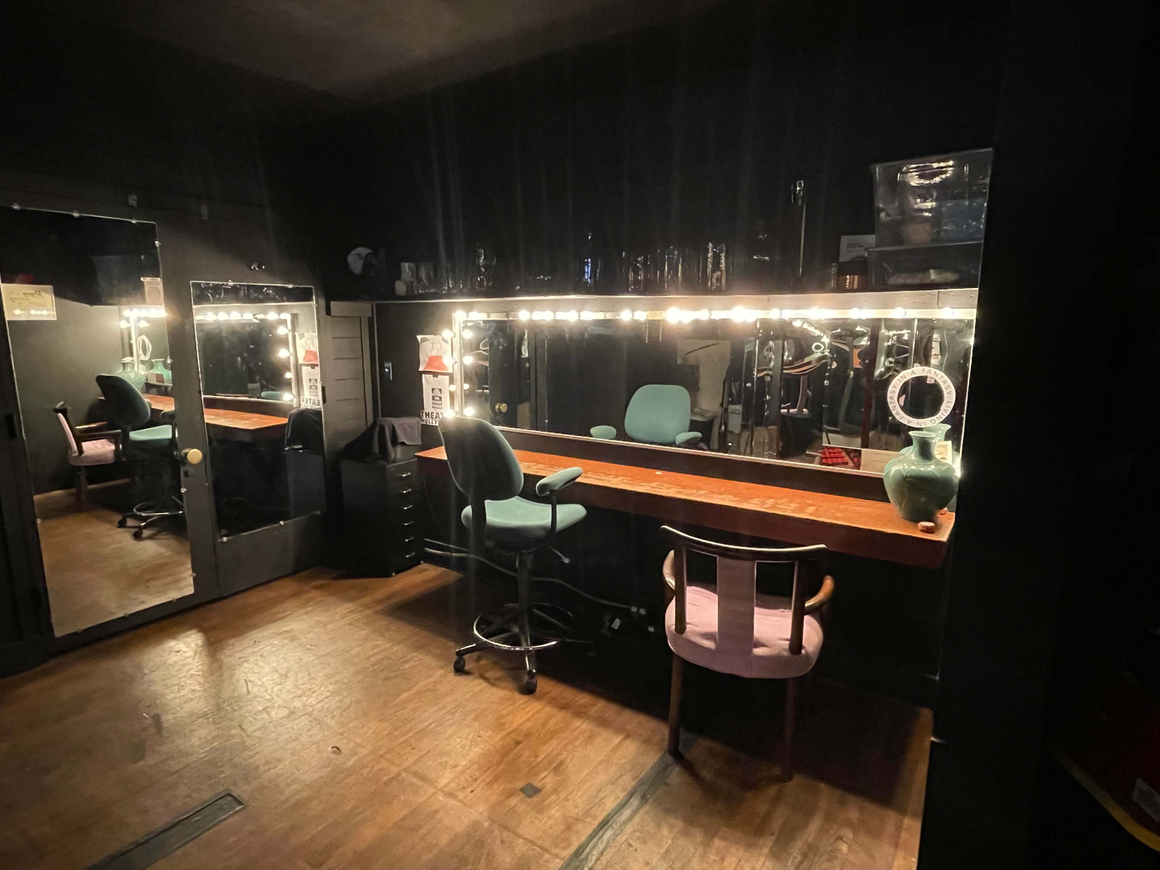 A dimly lit dressing room with a large mirror, illuminated by lights, and a wooden table surrounded by a chair and a stool.