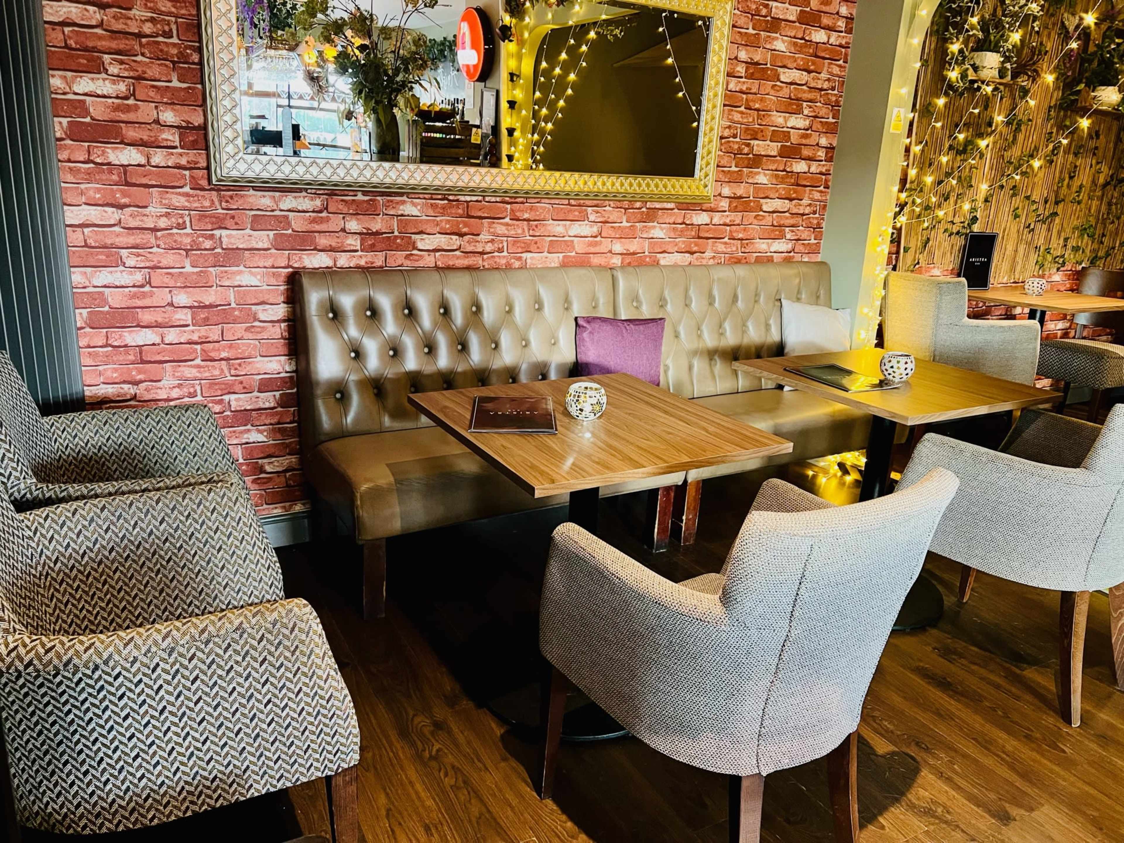 The image shows a cozy restaurant seating area with a brown tufted leather booth, wooden tables, and various patterned chairs against a brick wall.