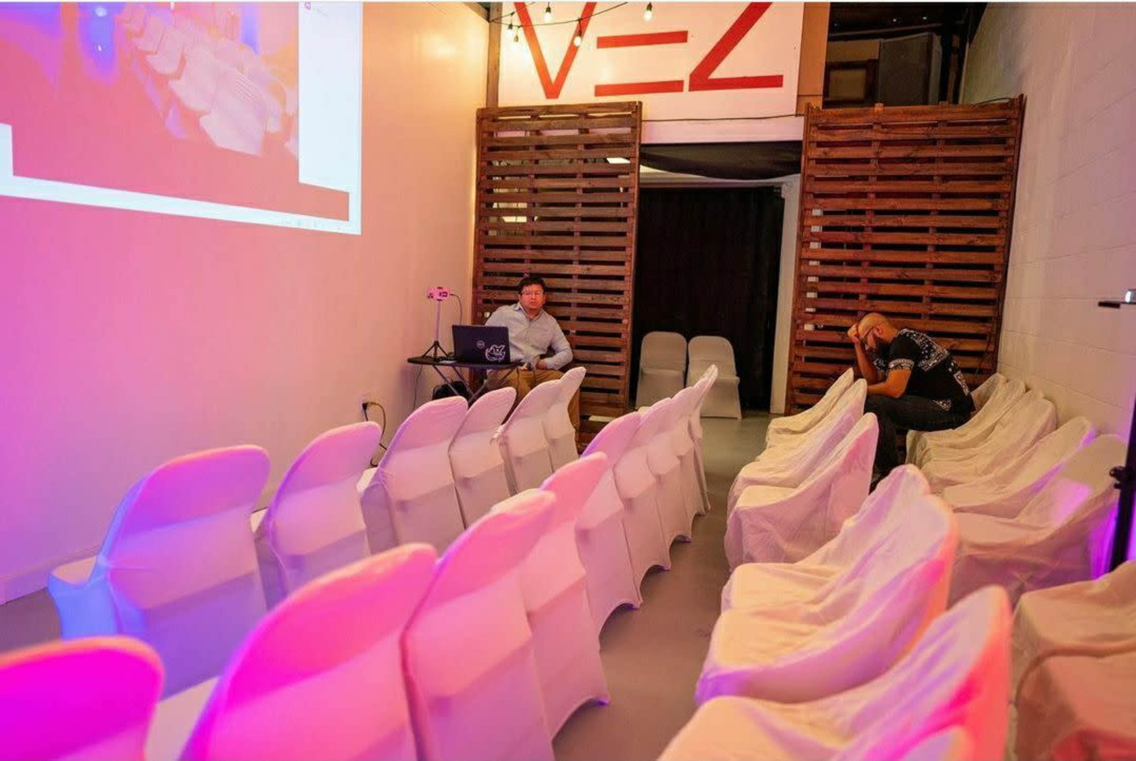 A partially filled room with rows of white chairs, a man sitting at a table with a laptop, and another man reclining against the wall in the background.