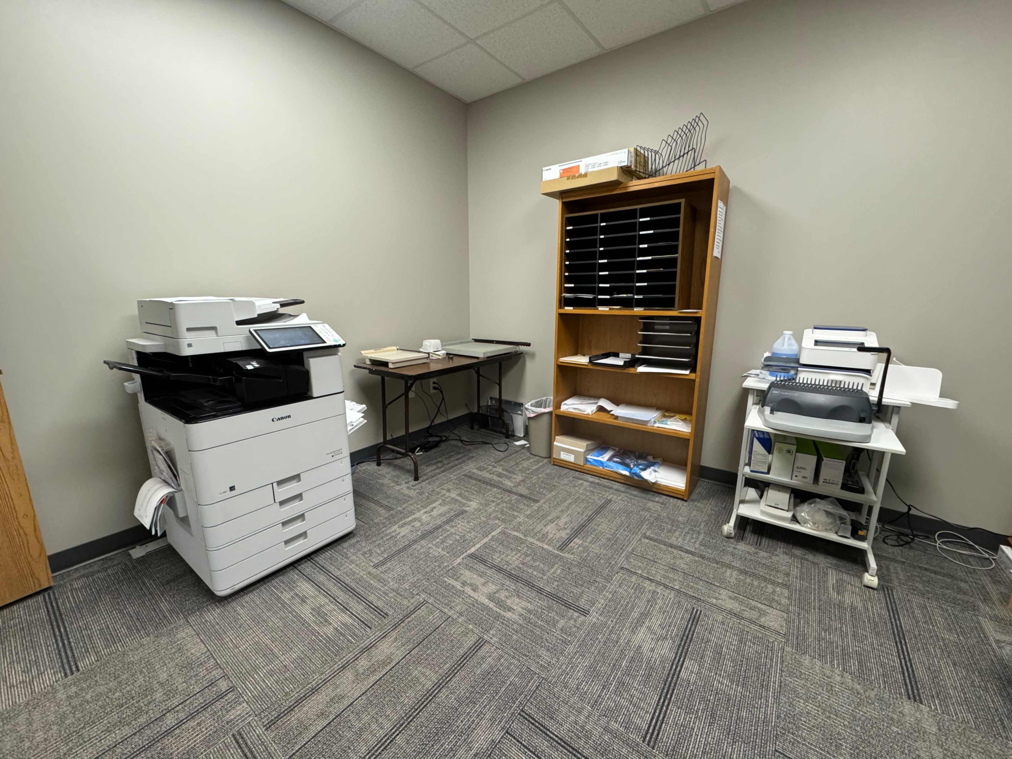 The image shows a small office space featuring a copier, a table, and a shelf with various office supplies.