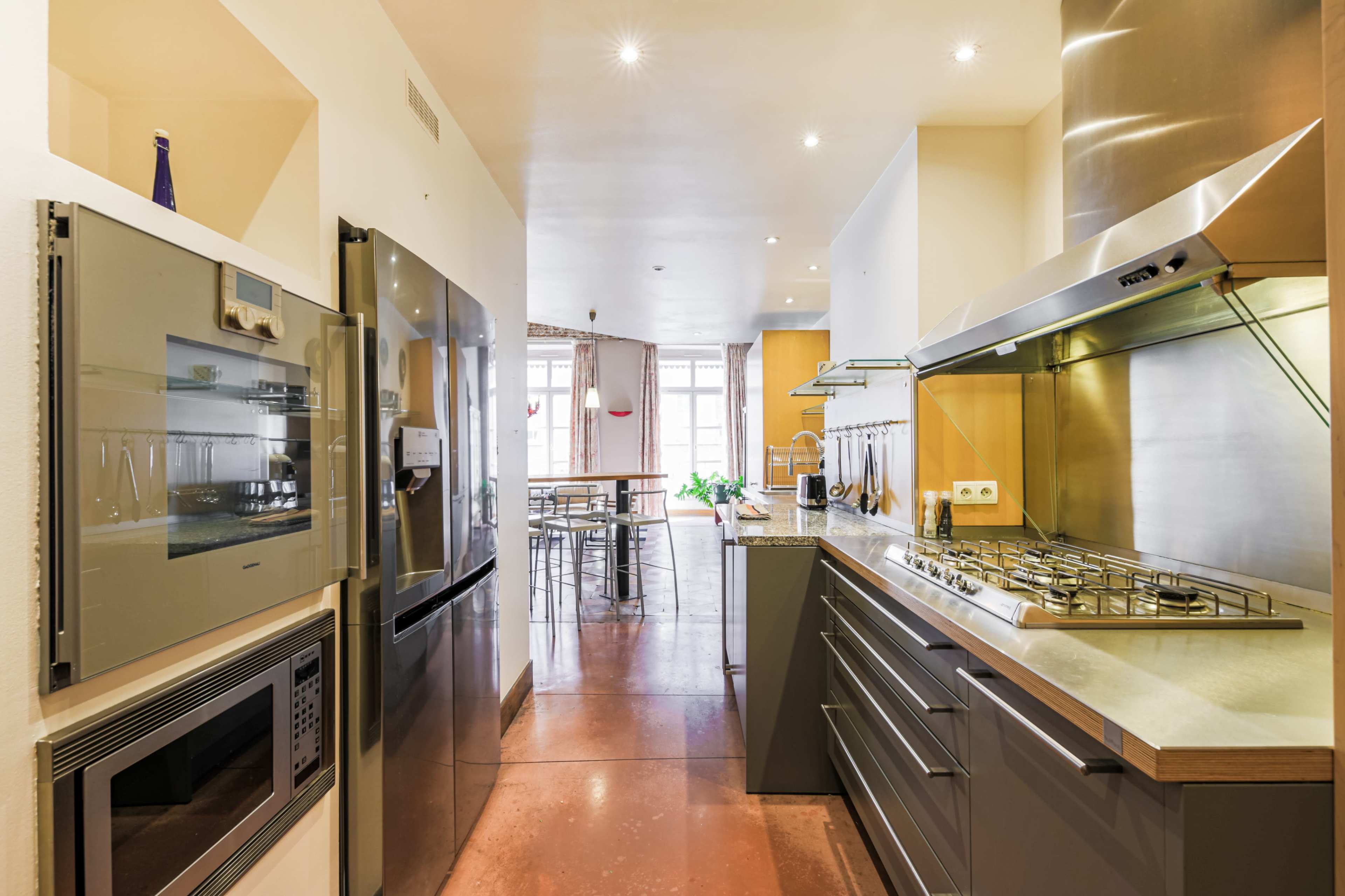 The image shows a modern kitchen with stainless steel appliances, including an oven and gas range, along a long counter with a view toward a dining area.