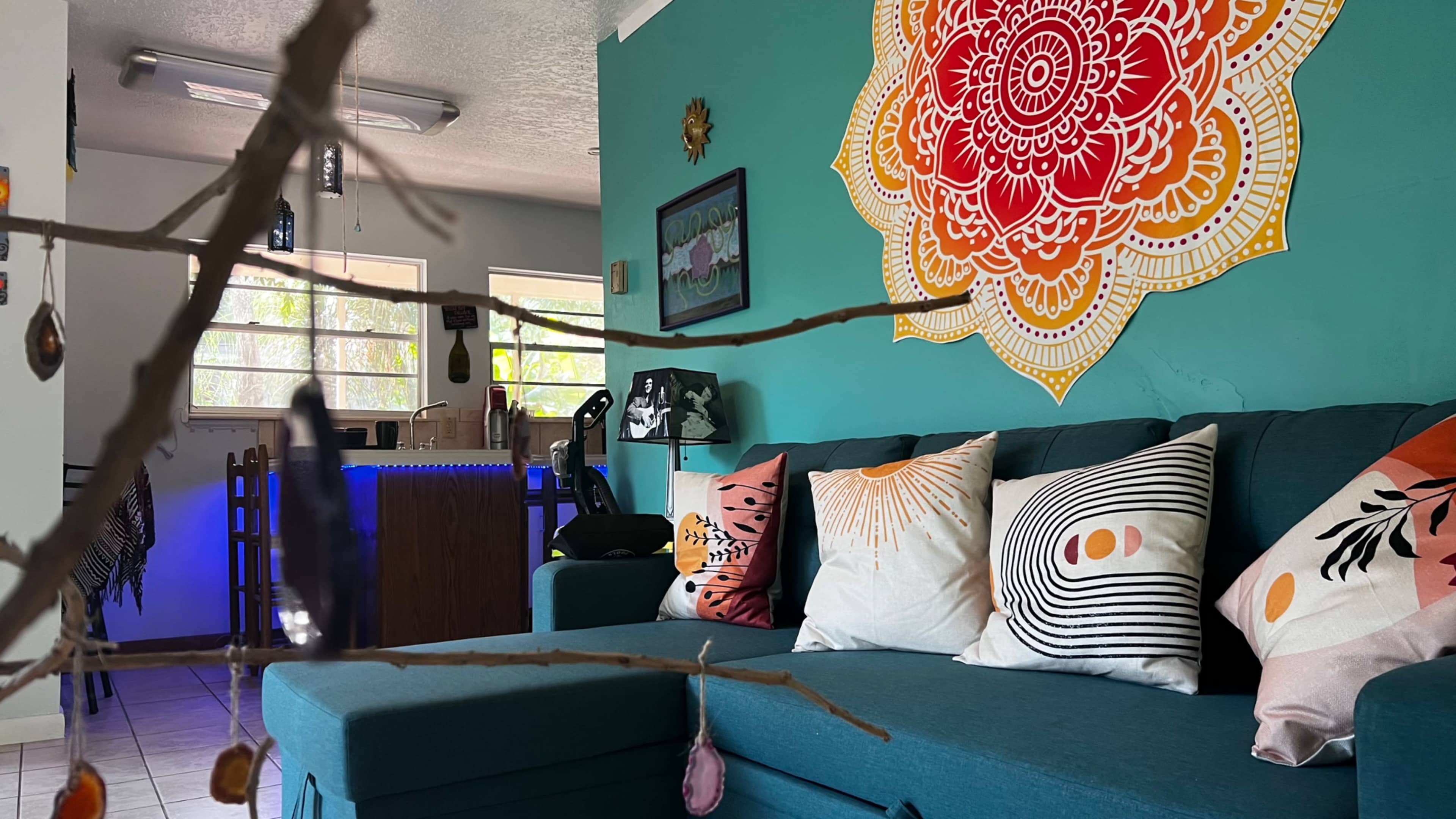 A teal living room features a large mandala artwork on the wall above a dark blue couch adorned with decorative pillows, with a branch hanging in the foreground.