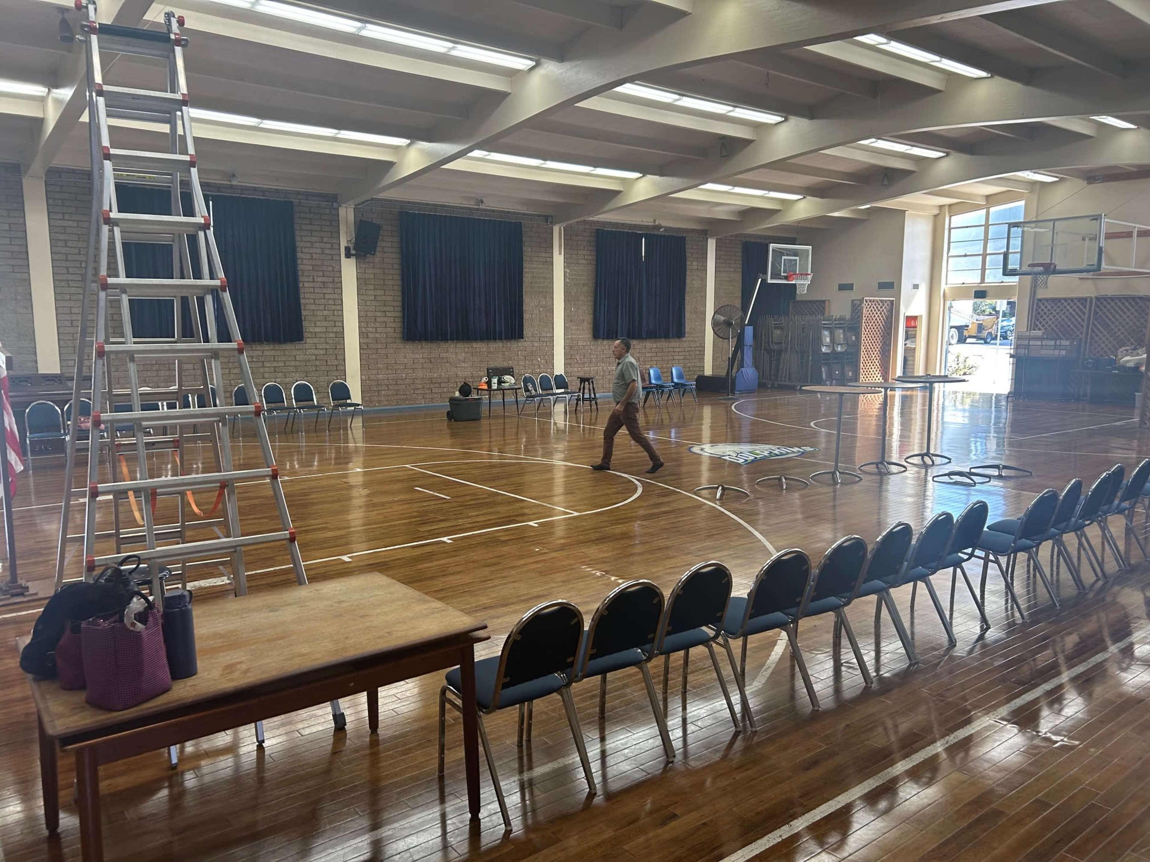 A person walks across a basketball court with chairs arranged around the perimeter and a ladder leaning against the wall.