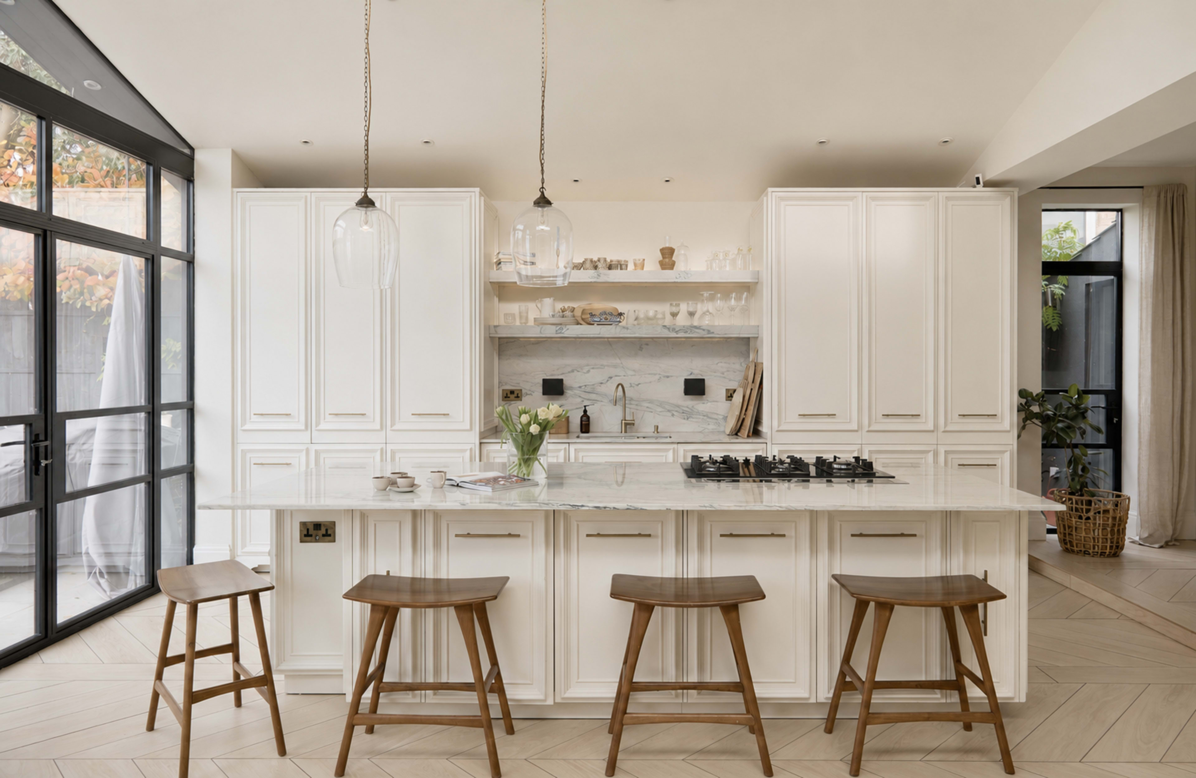 The image shows a modern kitchen with white cabinetry, a marble countertop, and three wooden stools at a central island.