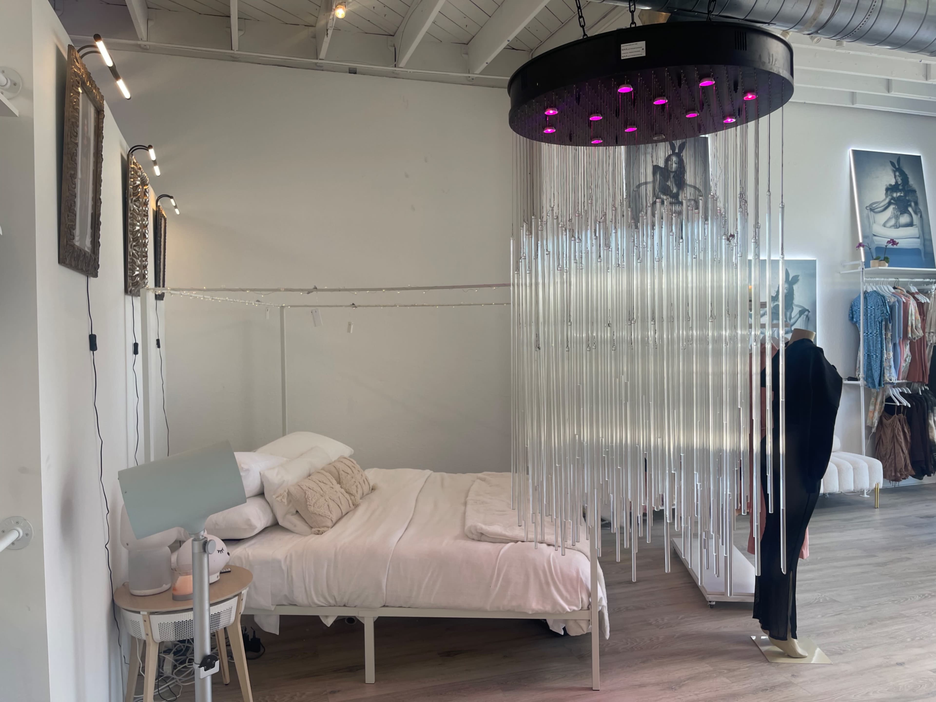 The image shows a bedroom setup featuring a bed with white bedding, a side lamp, and a decorative hanging installation made of clear vertical rods, alongside a clothing display on the right.