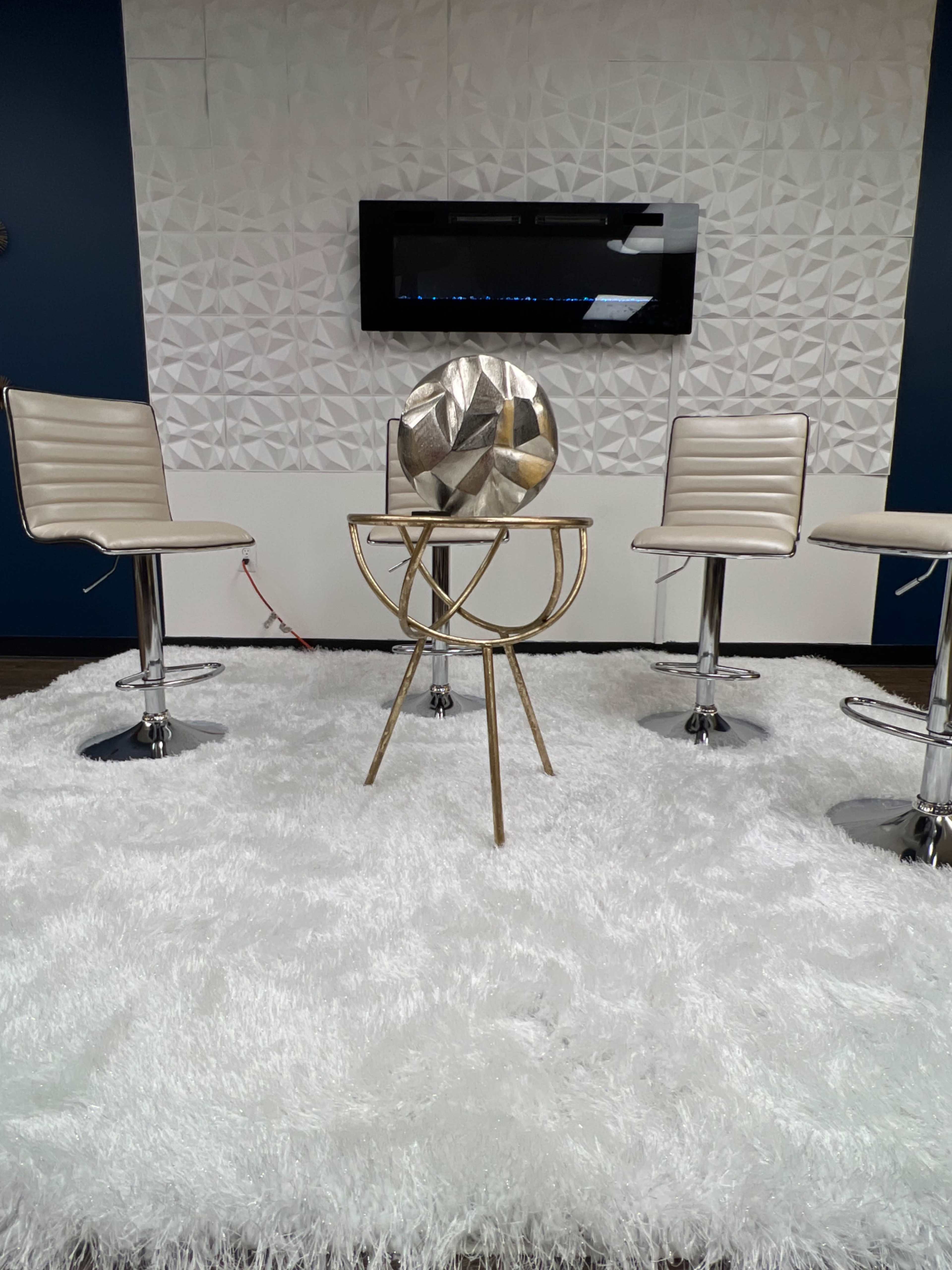 The image shows a modern seating area with three metal bar stools arranged around a round gold accent table on a white rug, against a textured white wall and a mounted electric fireplace.