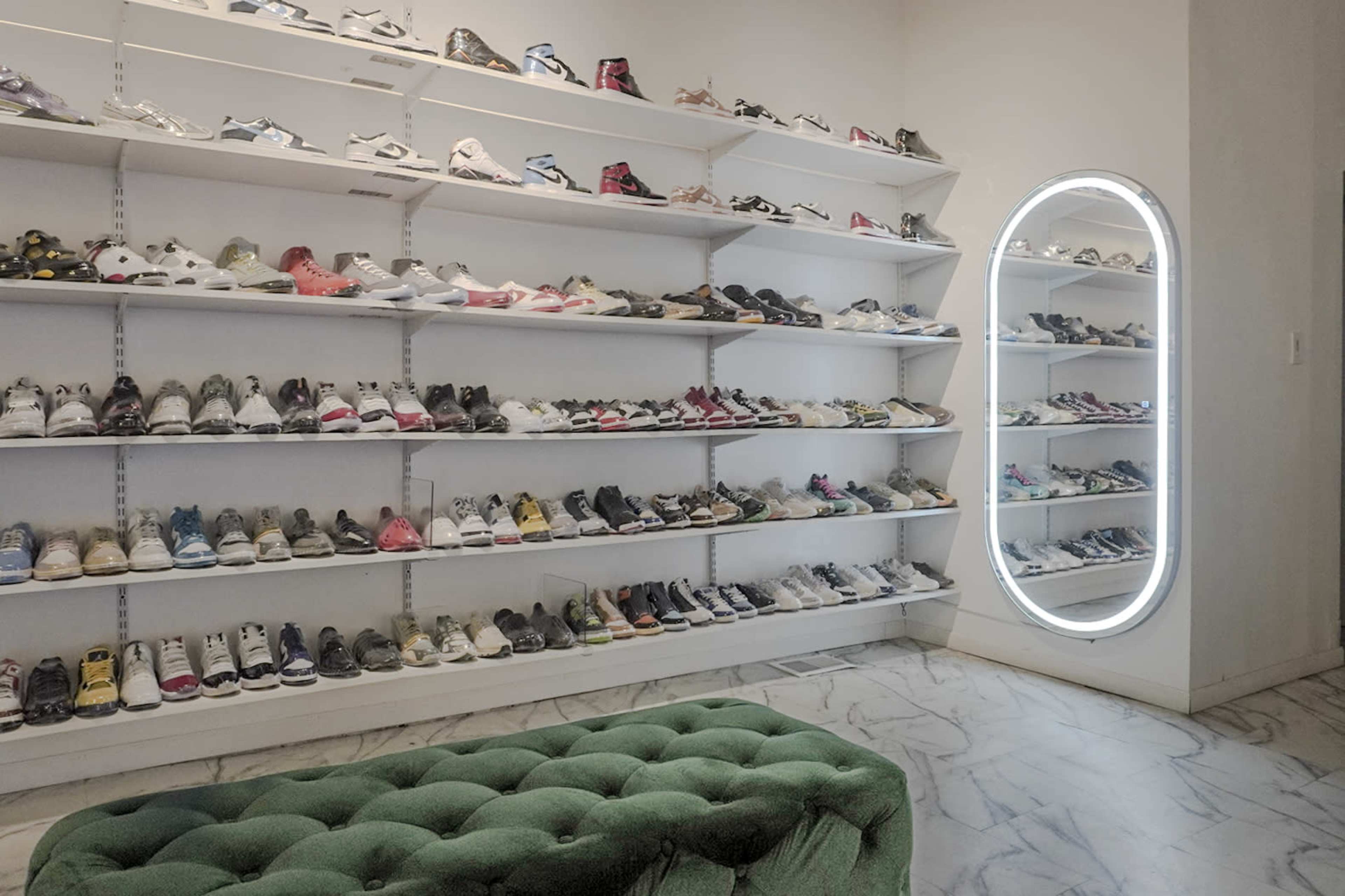 A neatly organized shoe wall displaying various styles of sneakers on shelves, alongside a round mirror and a tufted green bench in the corner.
