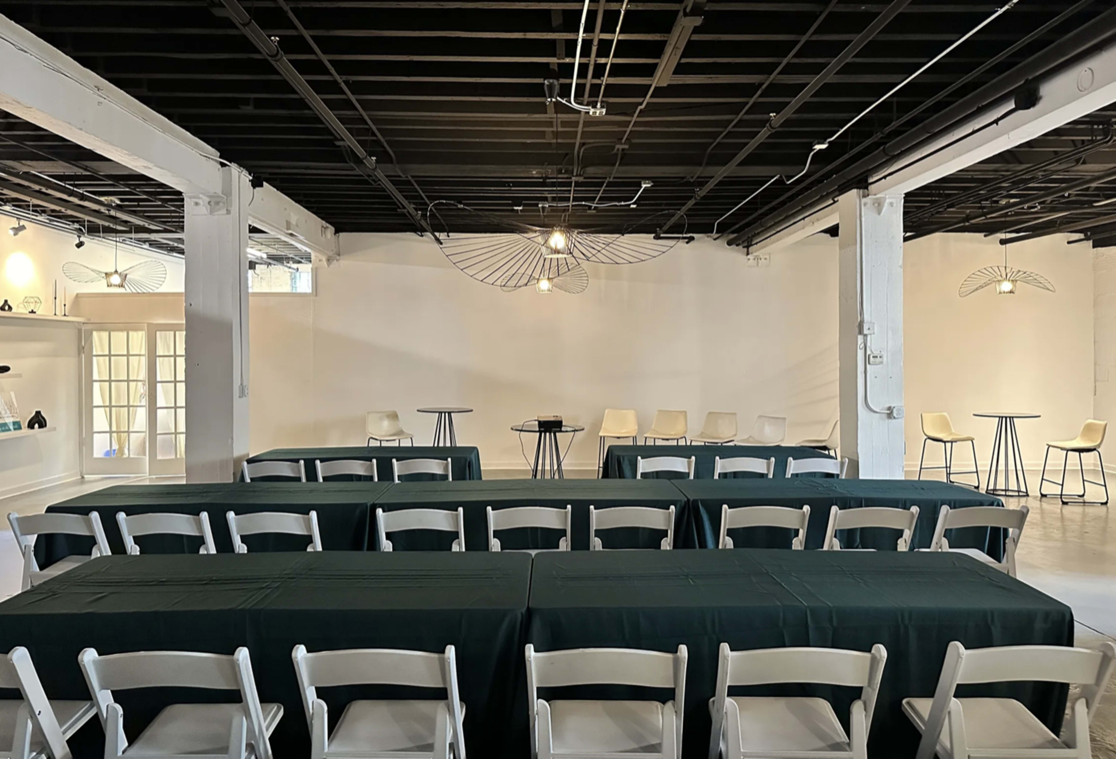 A spacious indoor venue arranged with green tablecloths covering long tables and a few high-top tables in a minimalist setting.