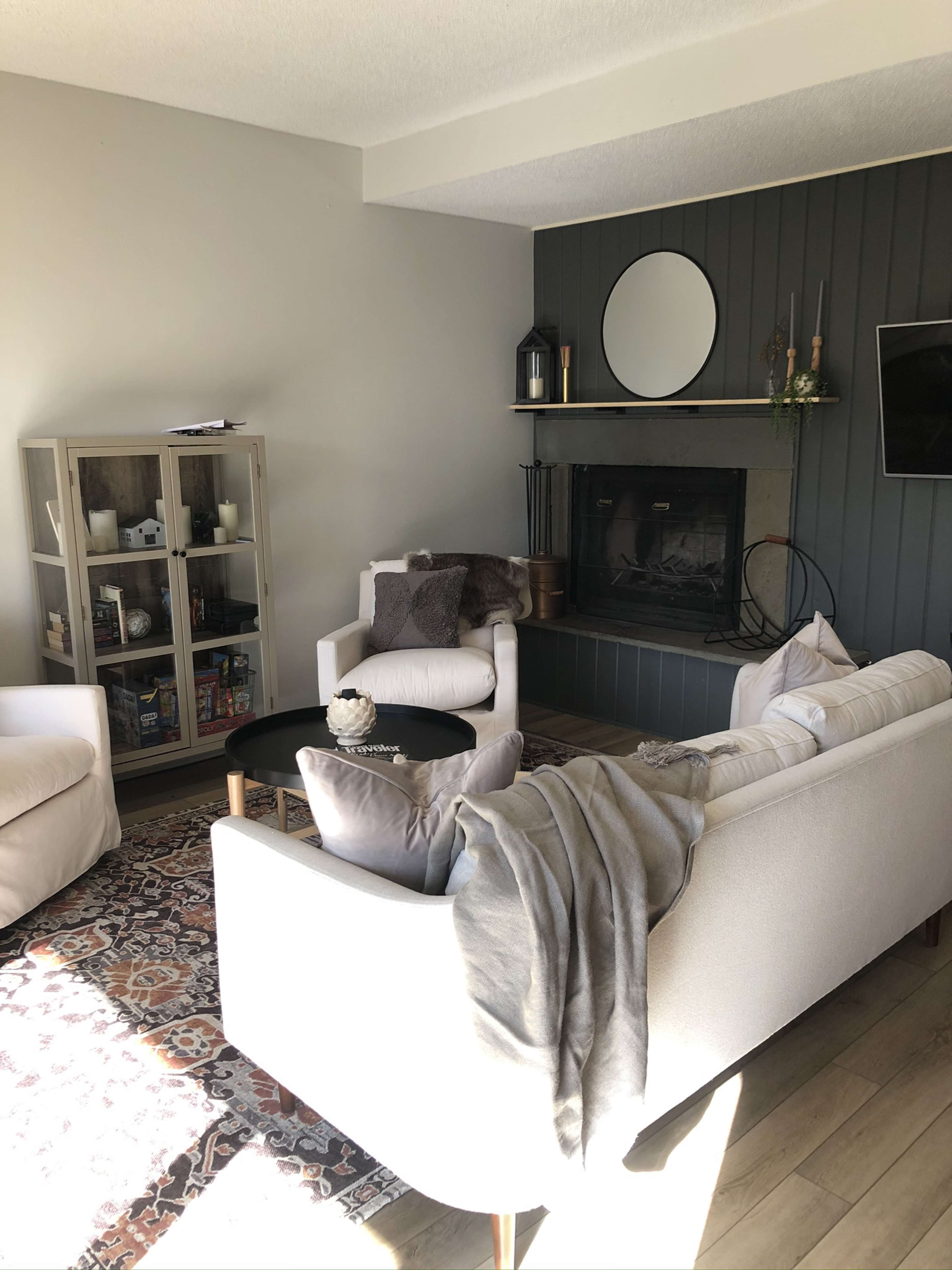 A cozy living room with a white sofa, two armchairs, a circular coffee table, and a fireplace, topped by a round mirror and decorated with plants.