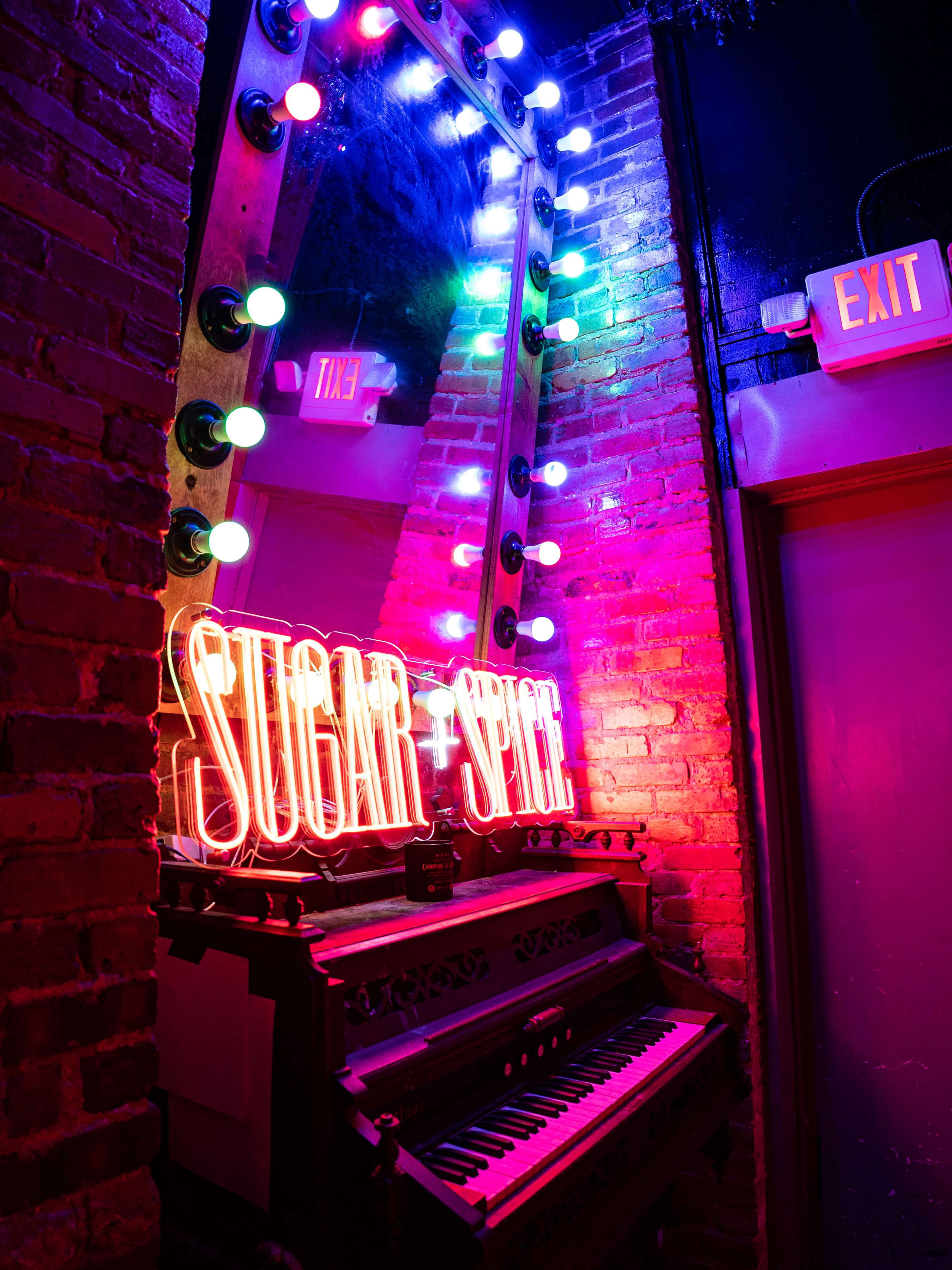 A brightly lit neon sign reading "SUGAR + SPICE" reflects in a mirror above an antique keyboard in a brick-walled space.