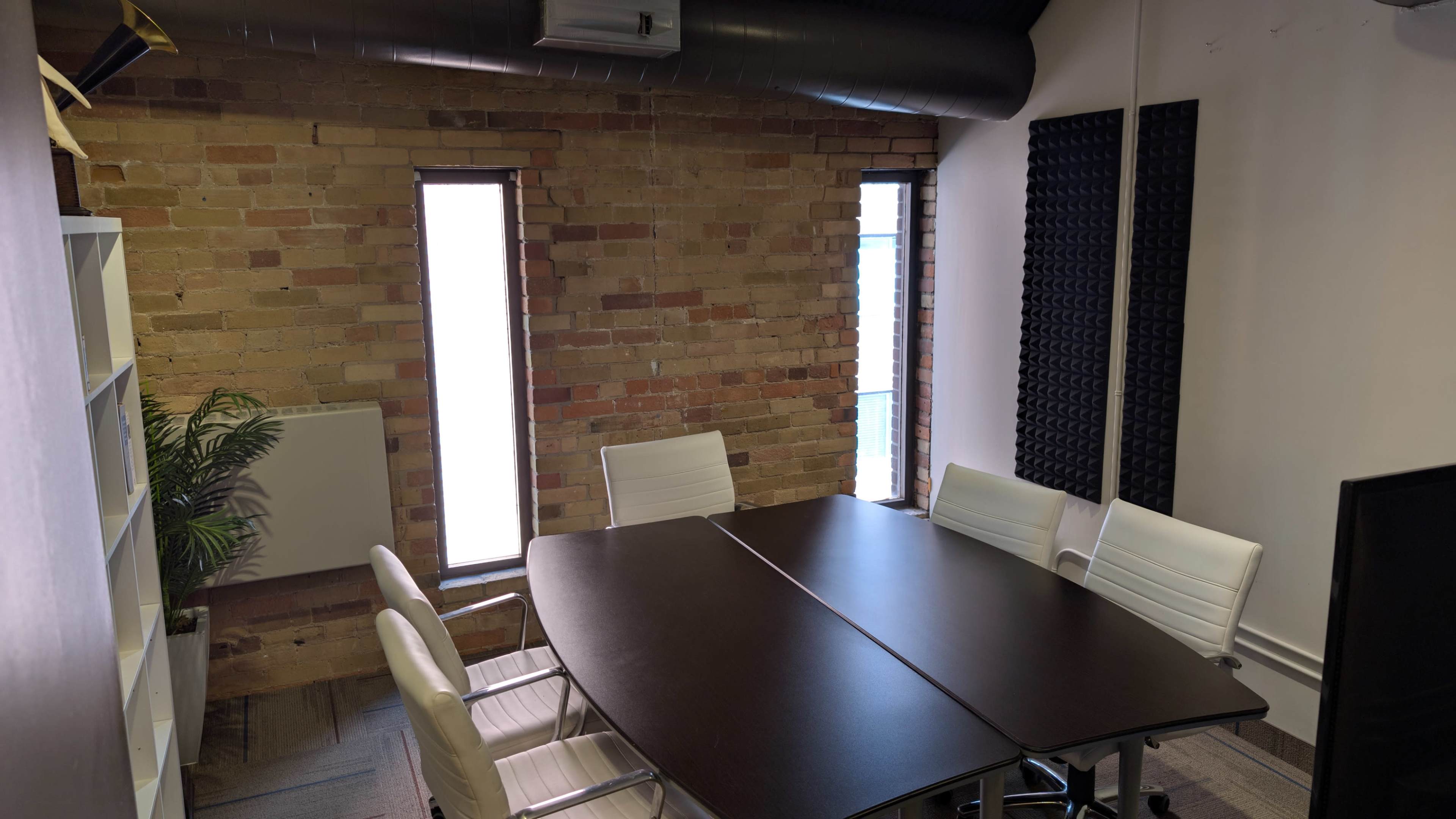 The image shows a meeting room with a dark wooden table surrounded by white chairs, exposed brick walls, and two windows.