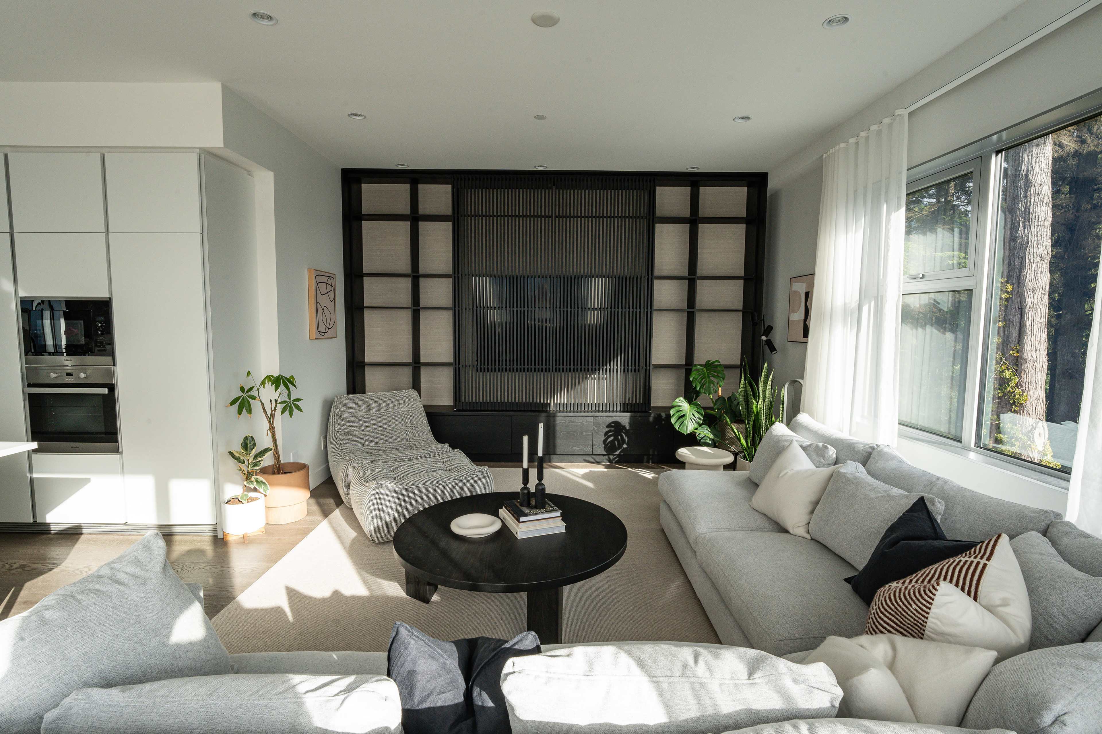 A spacious living room features a large sectional sofa, a round coffee table, a cozy lounging chair, and a wall-mounted television, surrounded by large windows and indoor plants.
