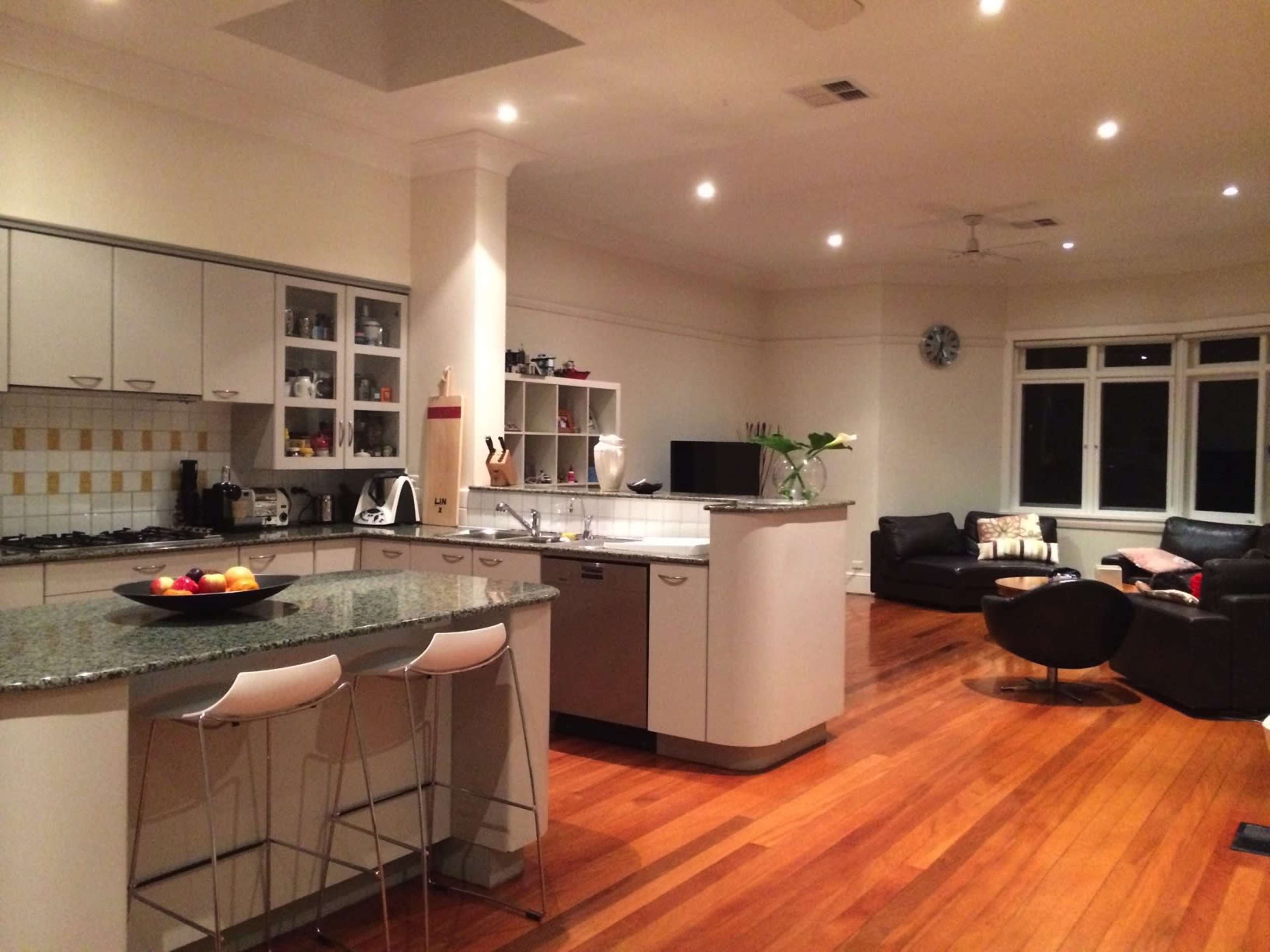 A modern kitchen with a large island featuring barstools, connected to a spacious living area with dark furniture and wooden flooring.