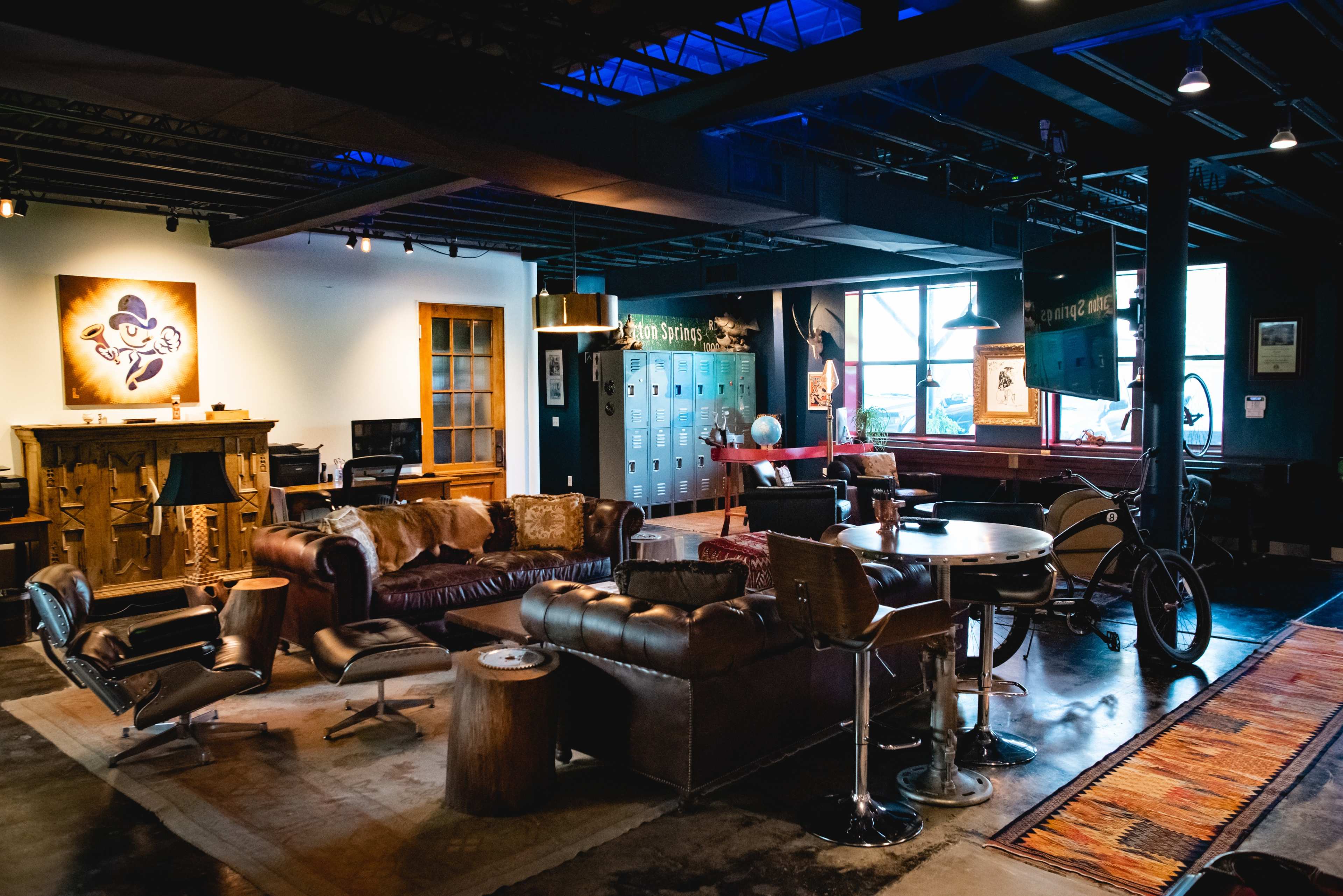 The image shows a spacious, industrial-style lounge area with leather seating, a wooden table, and various decorative elements, including lockers and art on the walls.
