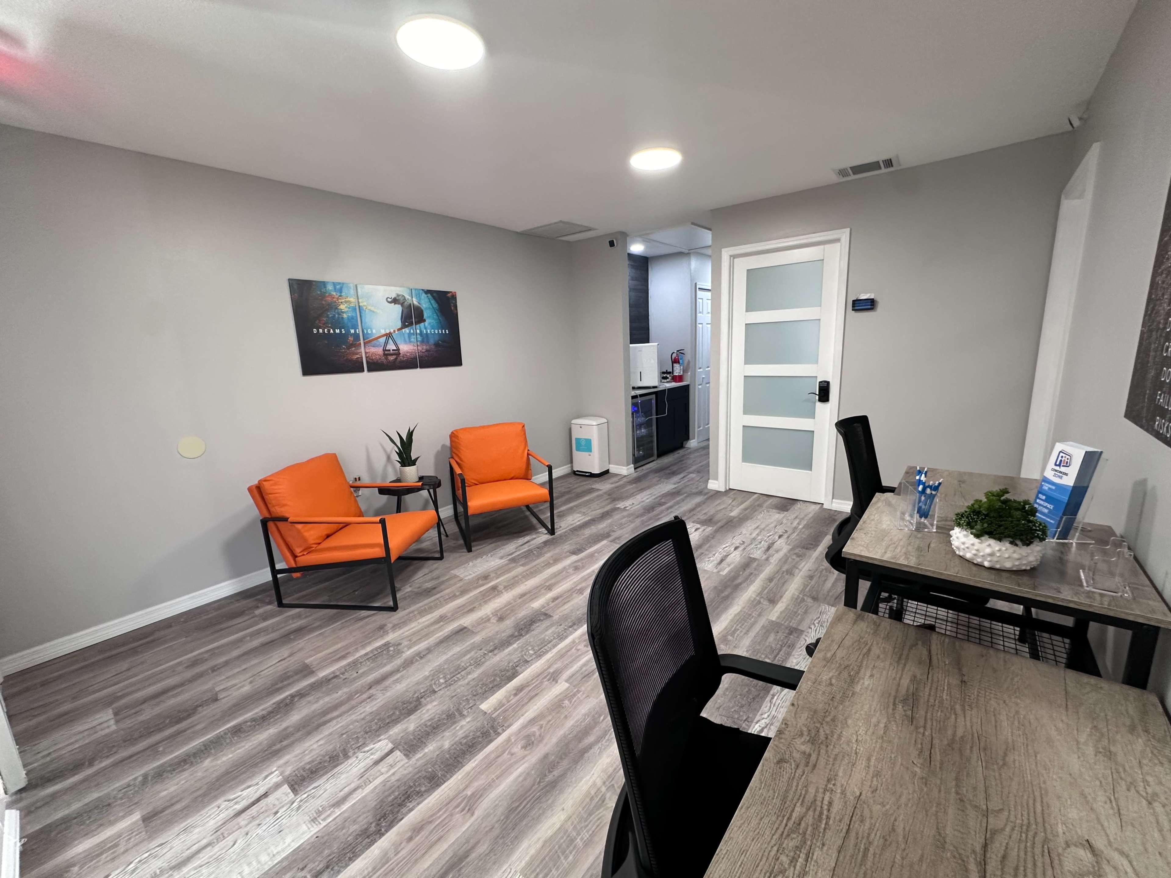 The image shows an office waiting area with two orange chairs, a desk with a plant, and a kitchenette area in the background.