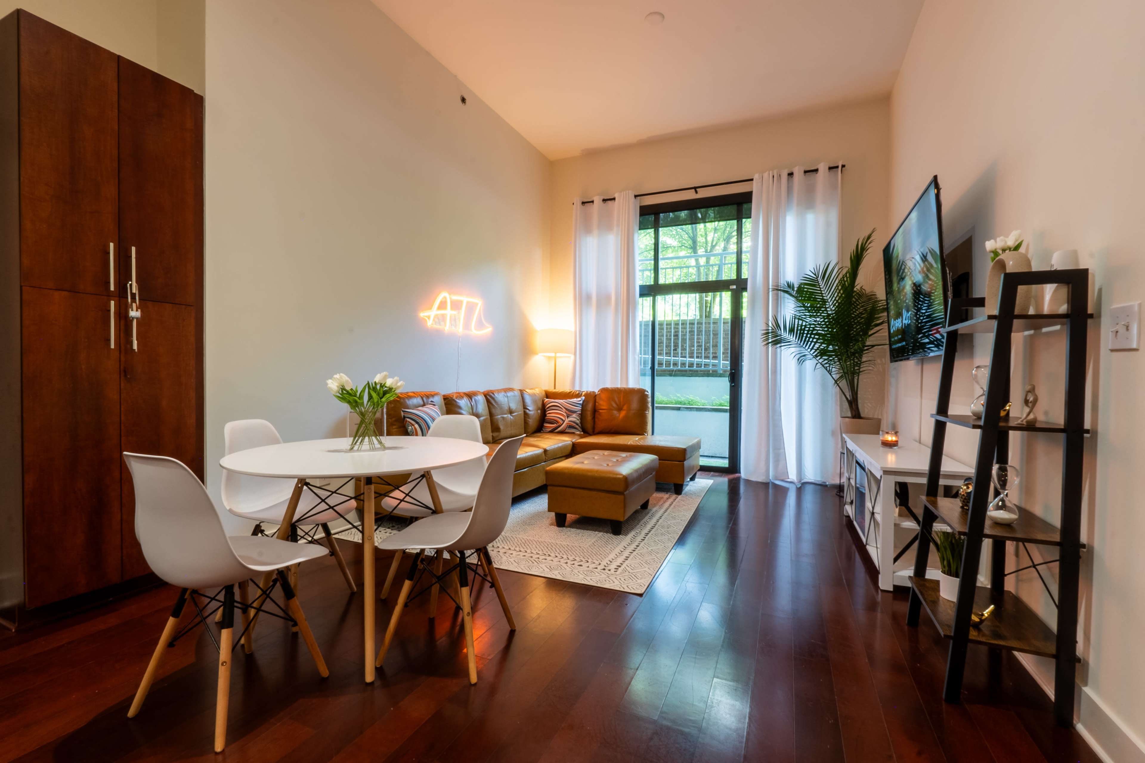 A modern living area featuring a brown leather sofa, a round dining table with white chairs, and a large window with sheer curtains that provides natural light.