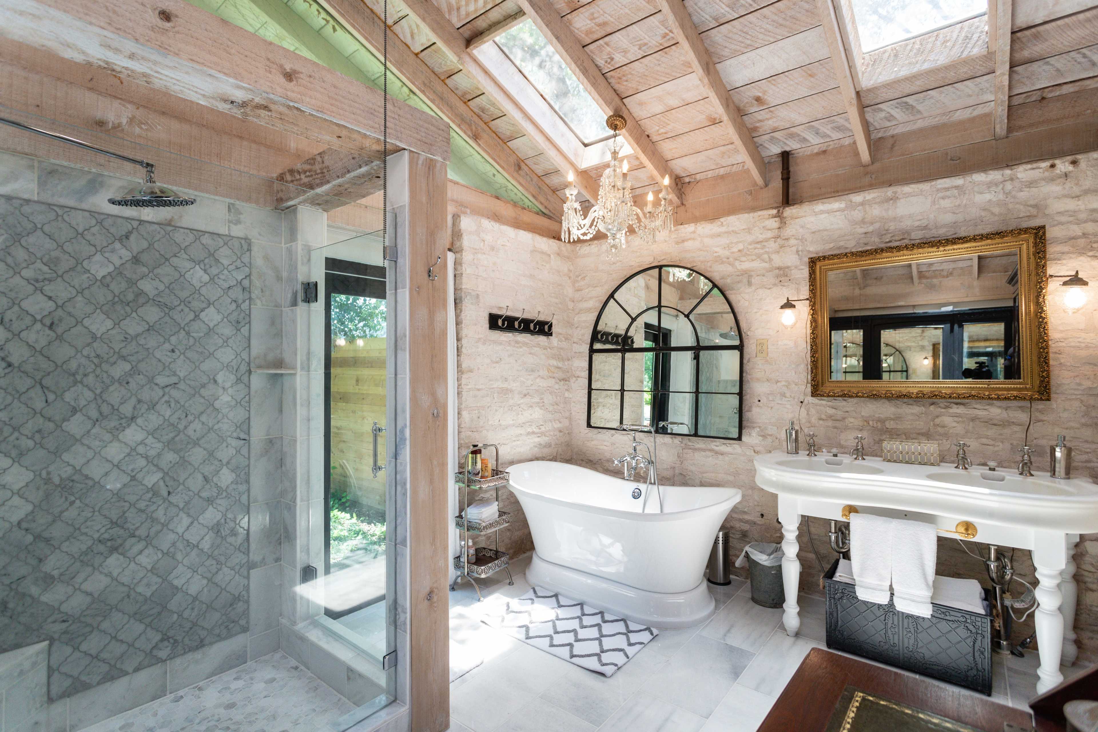The bathroom features a freestanding tub, a glass shower enclosure, and a large mirror with natural light streaming in from the skylights.