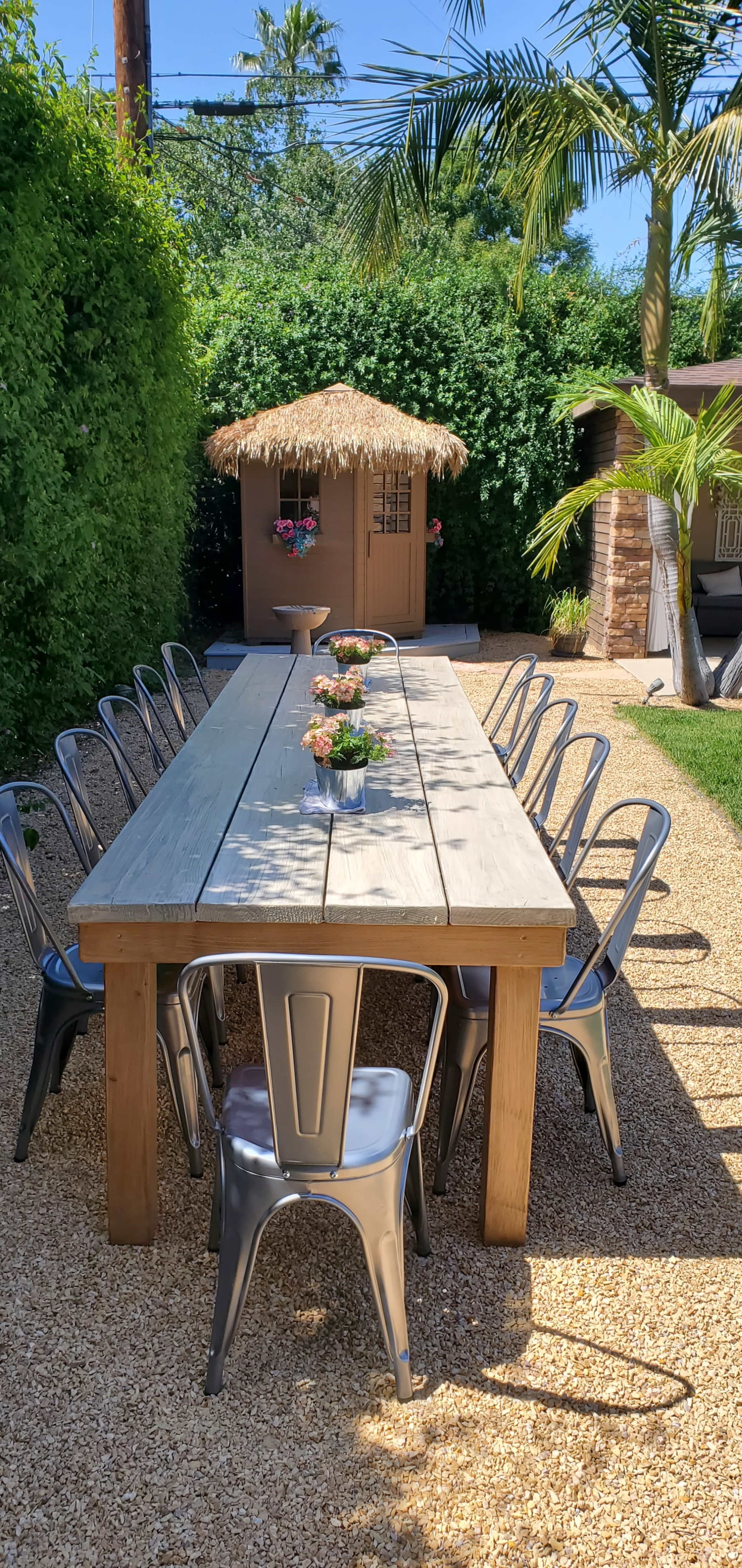 A long wooden dining table with metal chairs is set up in a landscaped backyard featuring a thatched-roof shed and lush greenery.