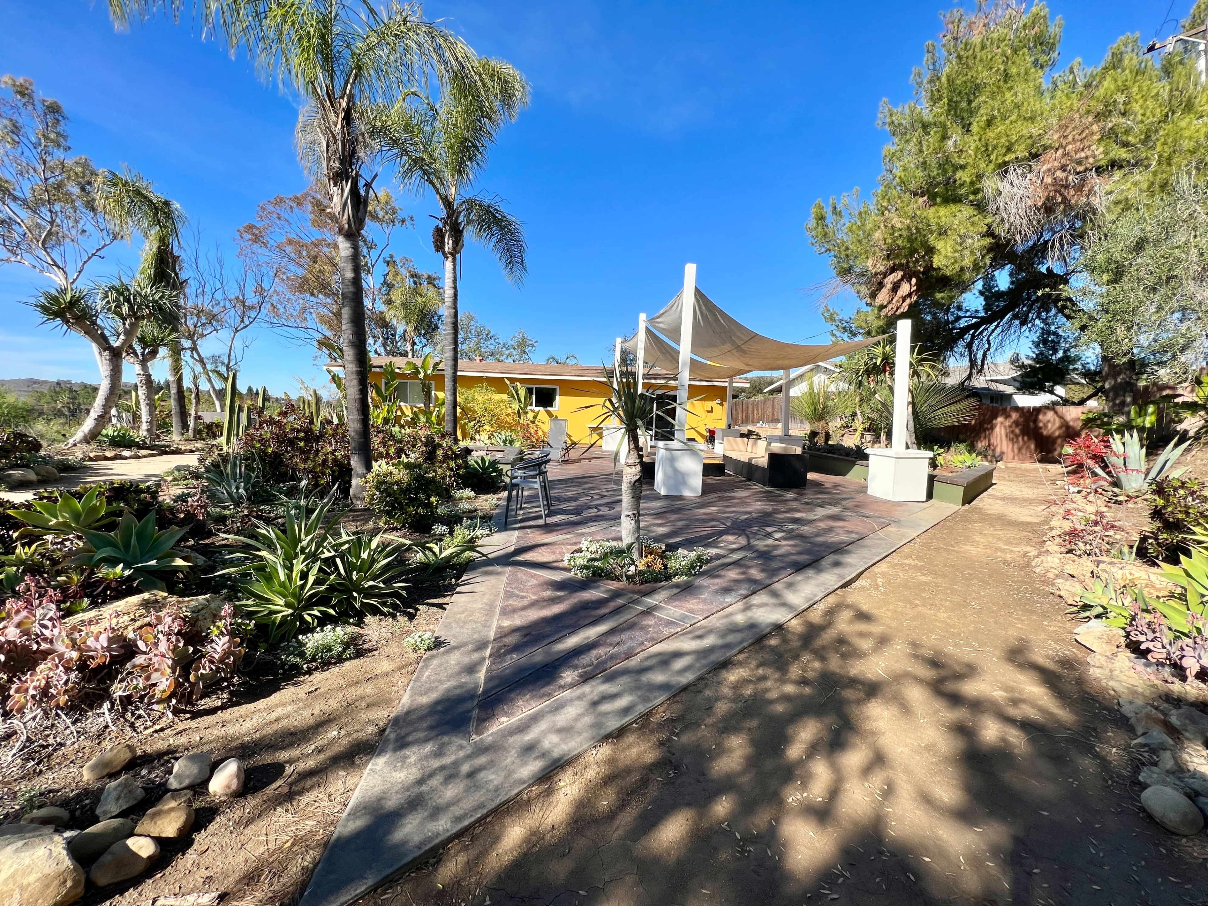 A landscaped outdoor area features a stone pathway leading to a covered seating space surrounded by various plants and trees.
