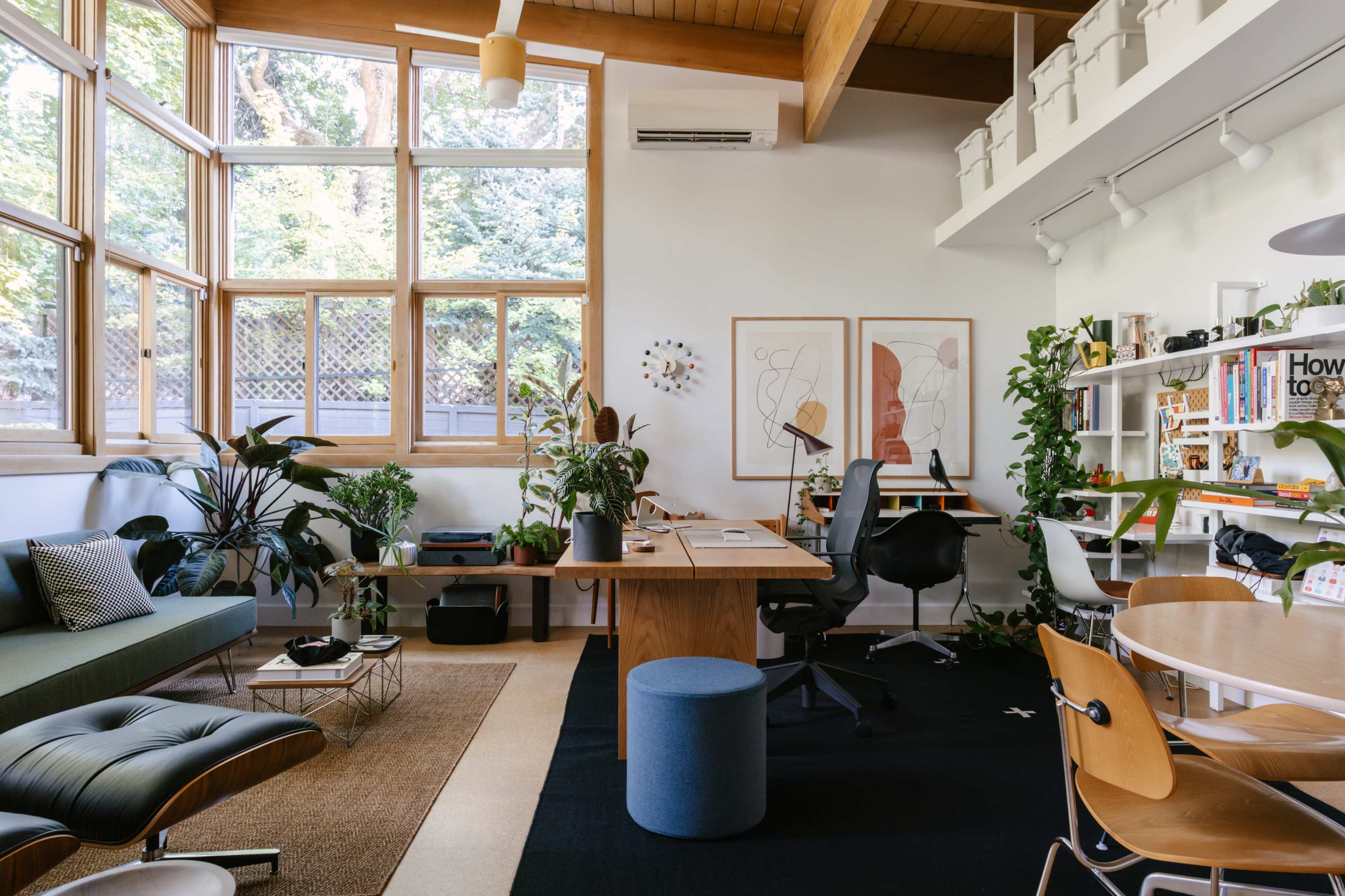 The image shows a home office with large windows, a wooden desk, a green sofa, plants, and a circular dining table surrounded by chairs.