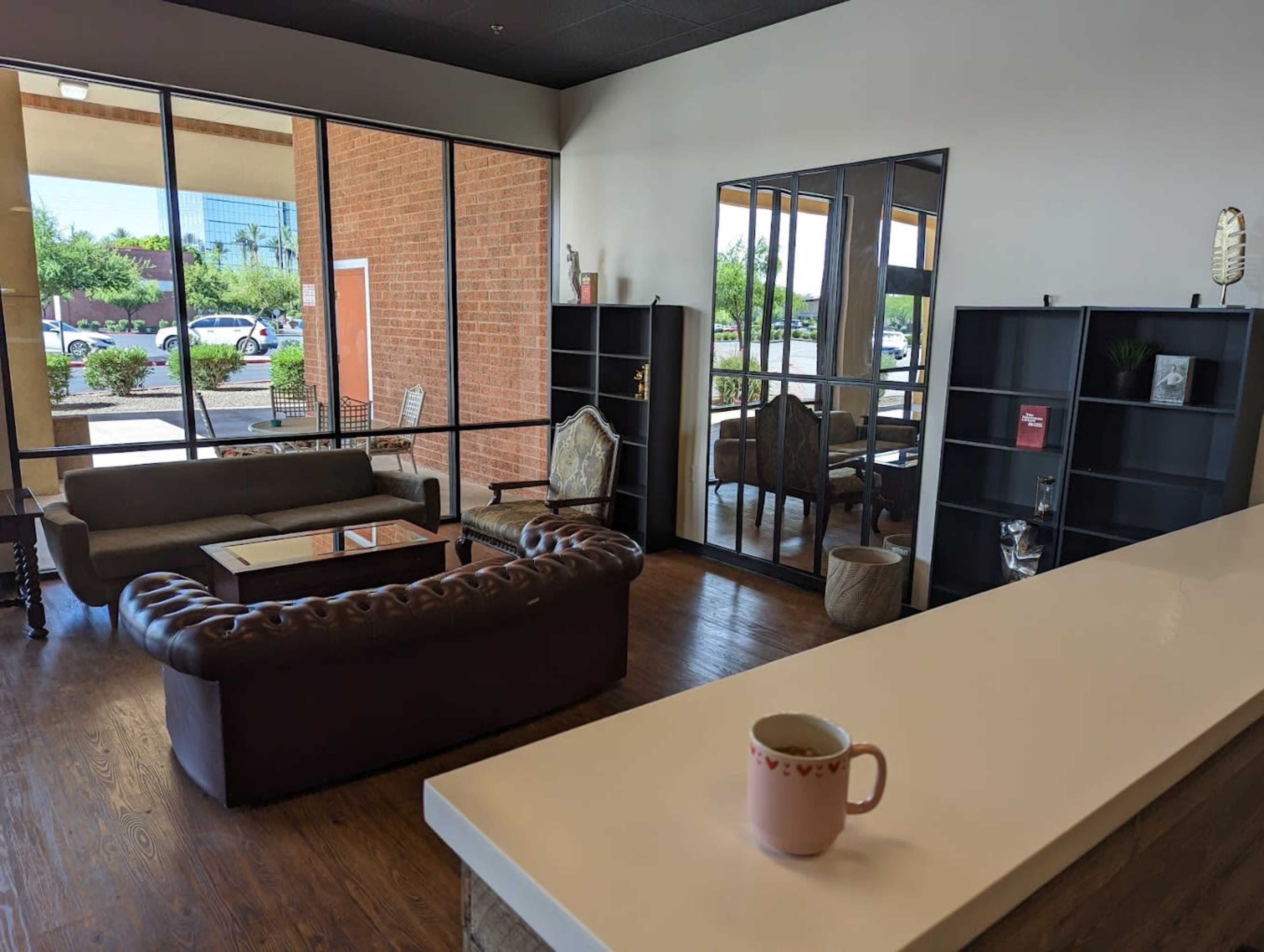 A modern waiting area featuring a brown leather sofa, a glass coffee table, a large mirror, and shelves, with large windows allowing natural light to enter.